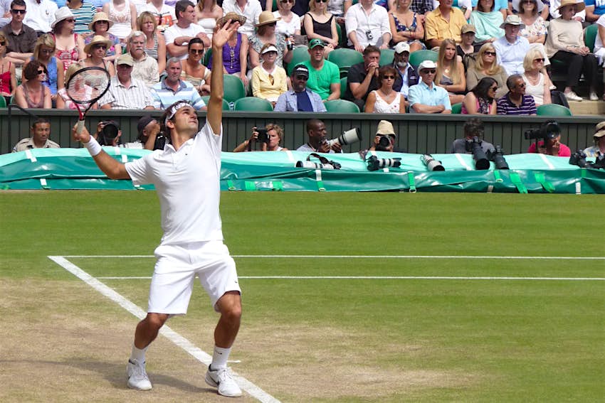 A close up of Roger Federer (dressed all in white) in the middle of his service motion; a colourful crowd behind him looks on