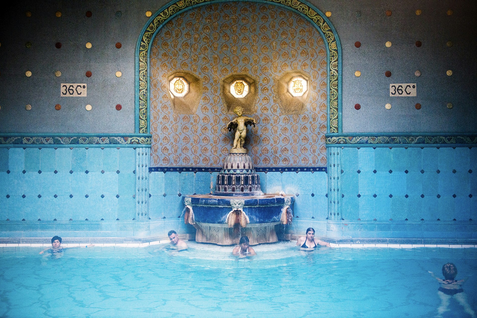 People sit in a steaming blue bath lined with blue tile next to an art nouveau fountain. A sign on the tiled wall indicates the water is 36C