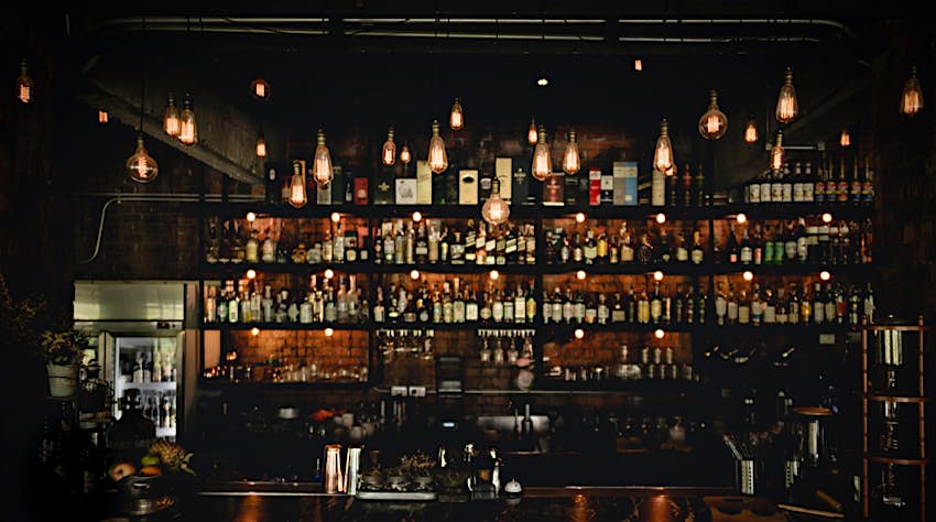 Shelves of bottles are illuminated by vintage lamps in the dark. There are glasses and other mix-drink instruments on the wooden bar. Cocktails can easily play a big role during a New Orleans weekend.     