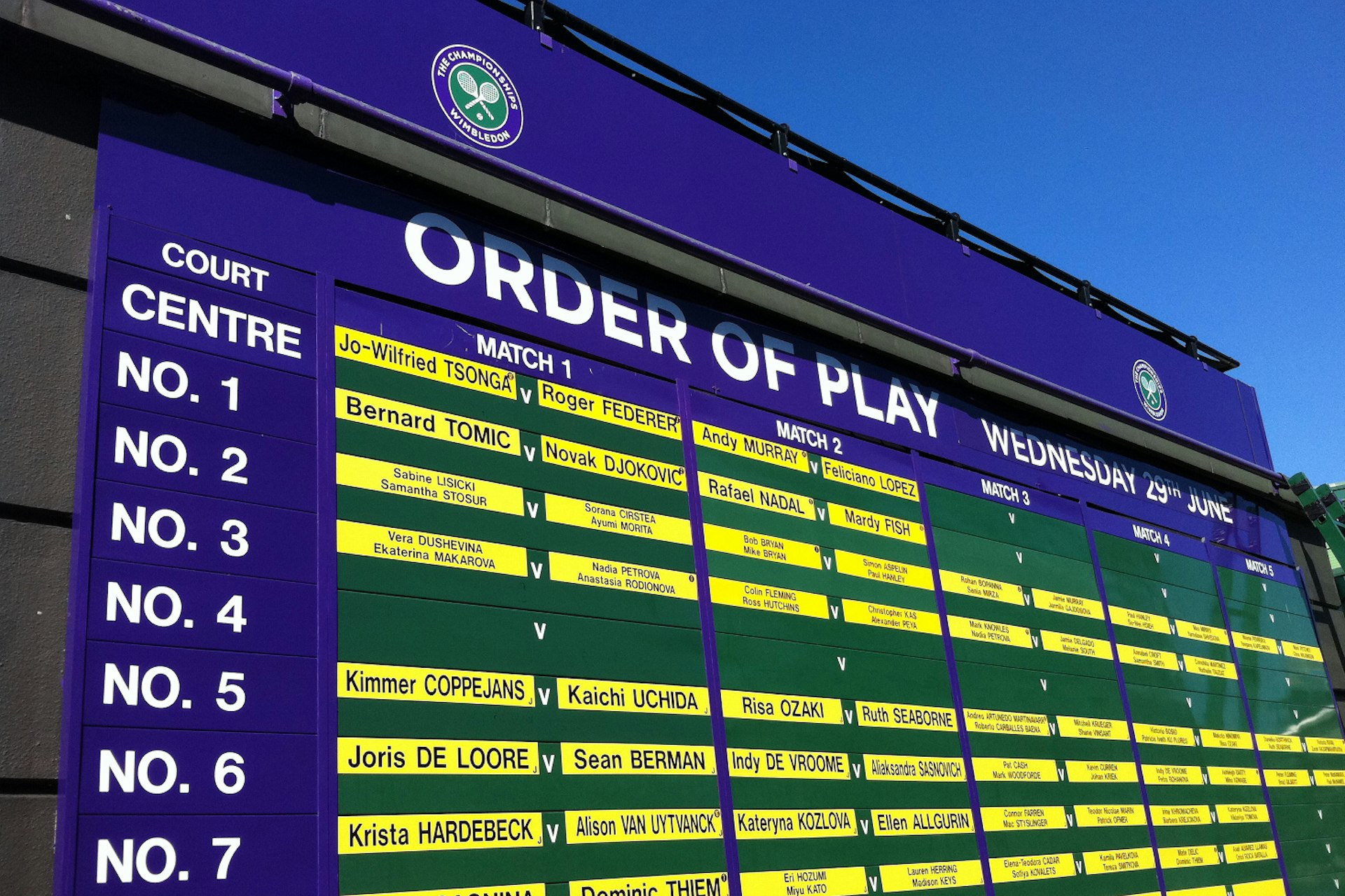 Under a blue sky sits a large purple and green billboard that notes (with bright yellow labels with black letters) what the first, second, third, fourth and fifth matches are on each court at Wimbledon ( noting the name of each competitor) on the day