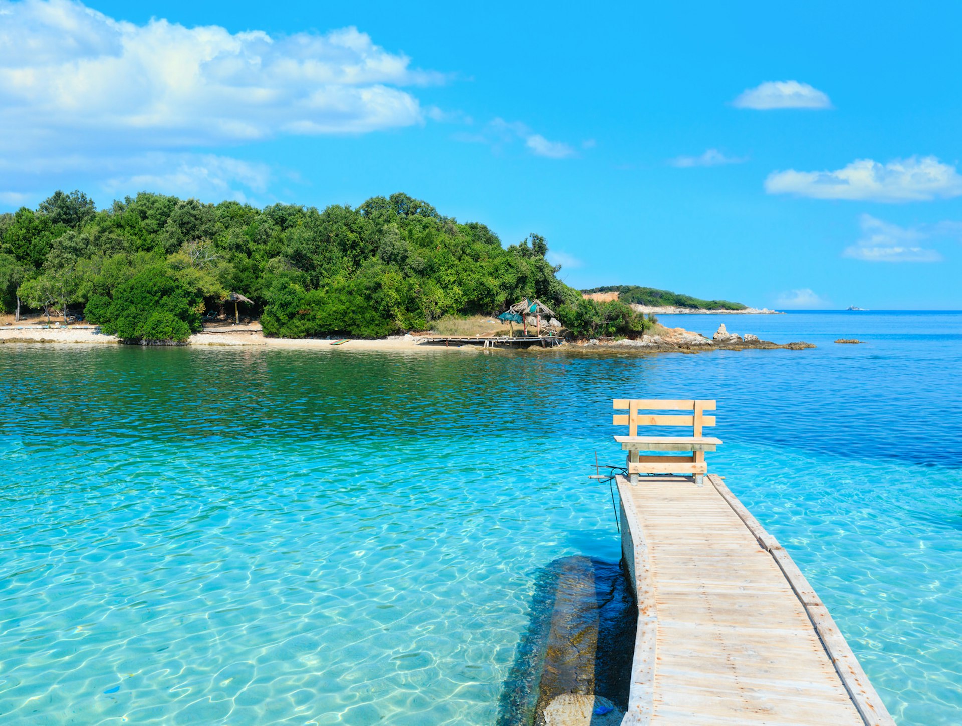 A wooden pier with a bench at the end stretches into shallow blue waters off the coast of Ksamil beach, with a small island, covered in trees, in the distance