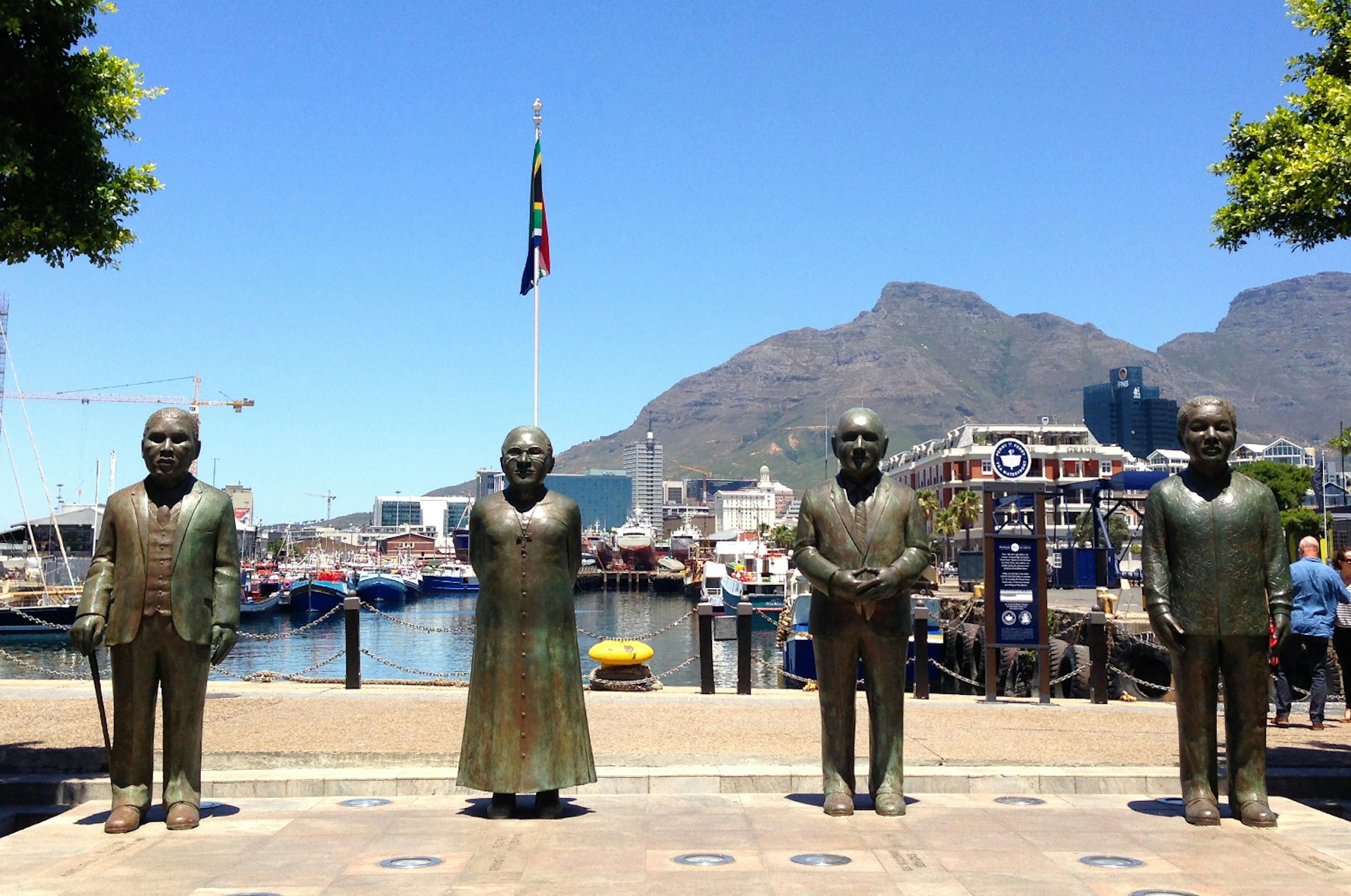 With Table Mountain as a backdrop, four bronze statues stand side-by-side in front of the V&A Waterfront and a flagpole with the South African flag.