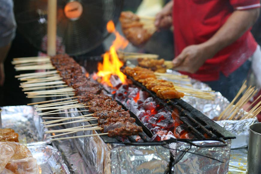 A row of satay skewers grilling over flaming coals © simonharrycollins / Shutterstock