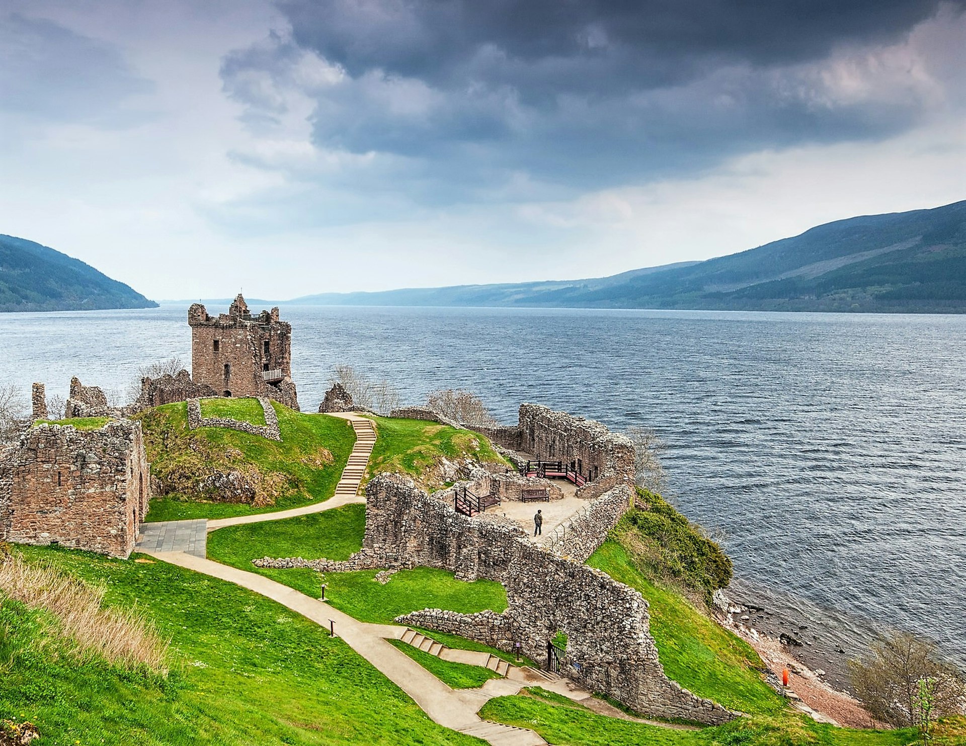 A castle on a green outcropping with a long winding road overlooking the dark blue expanse of Loch Ness