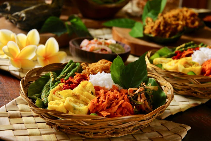 A popular Balinese meal of rice with variety of vegetables in a wooden bowls sitting on a bamboo place mats. Flowers are placed around the bowls of food. Dining in Bali can sometimes look like a work of art. 