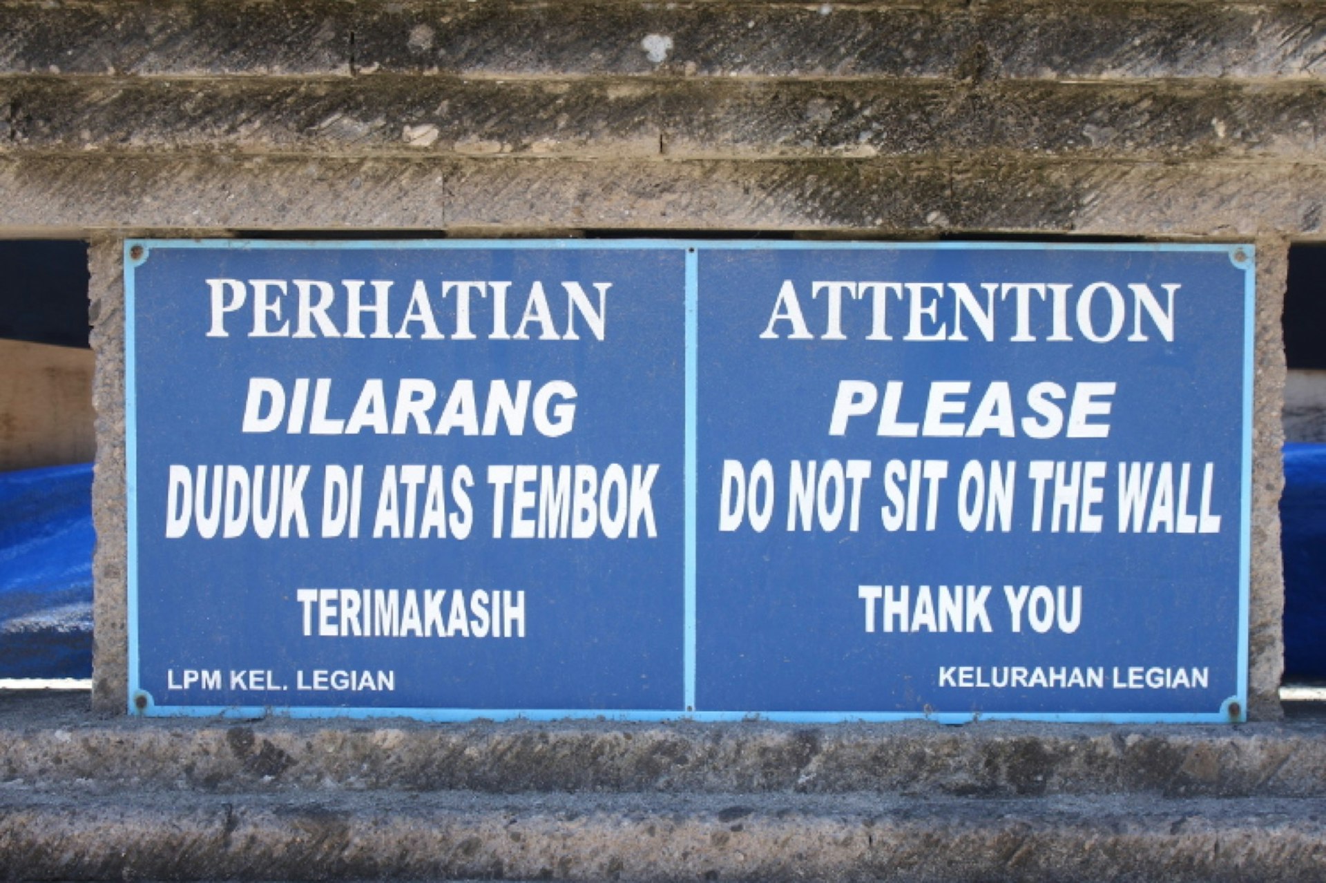 A pair of signs, one in Indonesian and the other in English, warning travellers to not sit on a wall in Bali. 