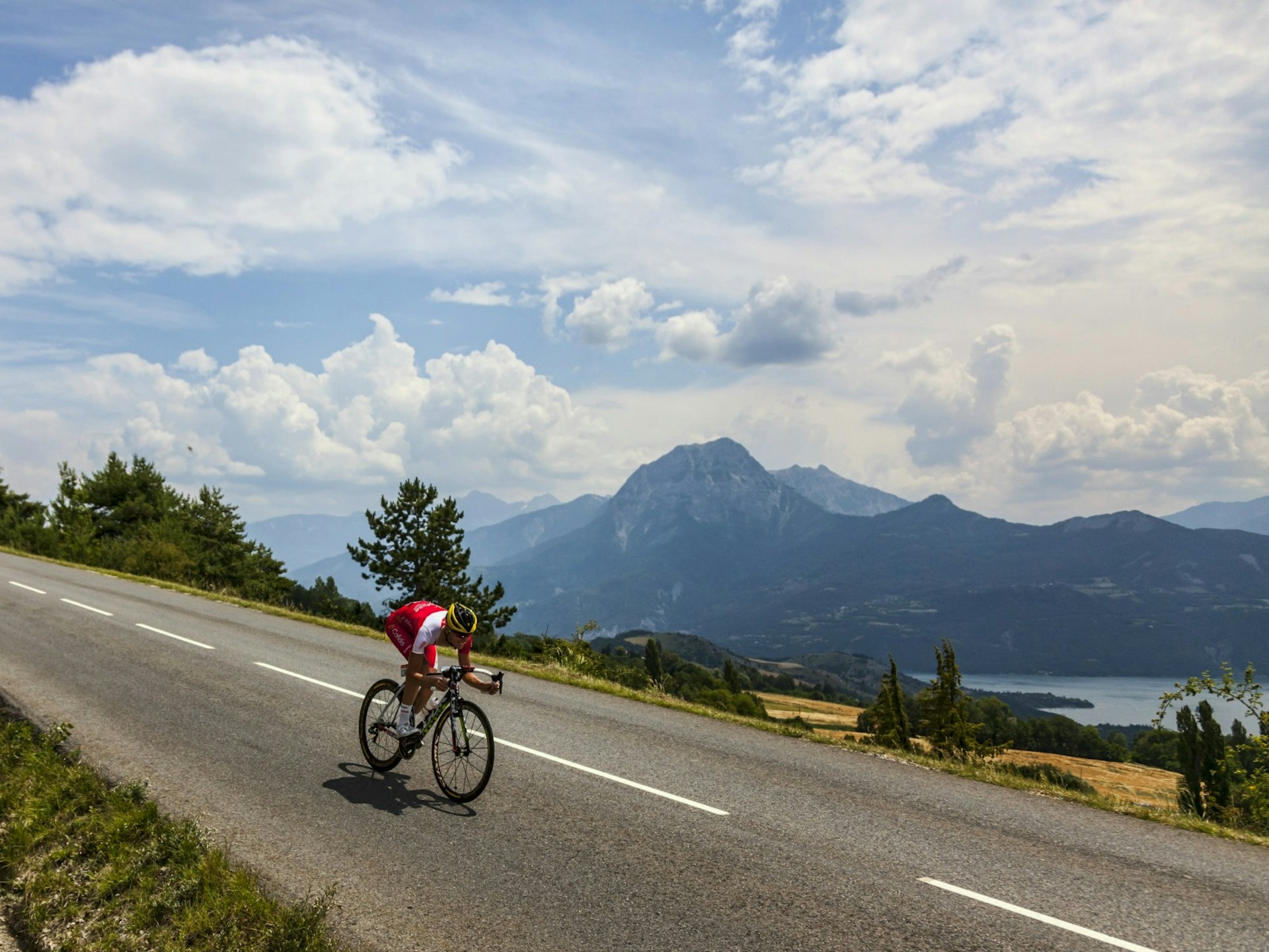 Tour de France - A cyclist rides between Embrun and Chorges during the Tour de France against a stunning backdrop of lakes, mountains and a cloud-streaked sky. The cyclist's jersey is red and white and they wear a sleek black and yellow helmet