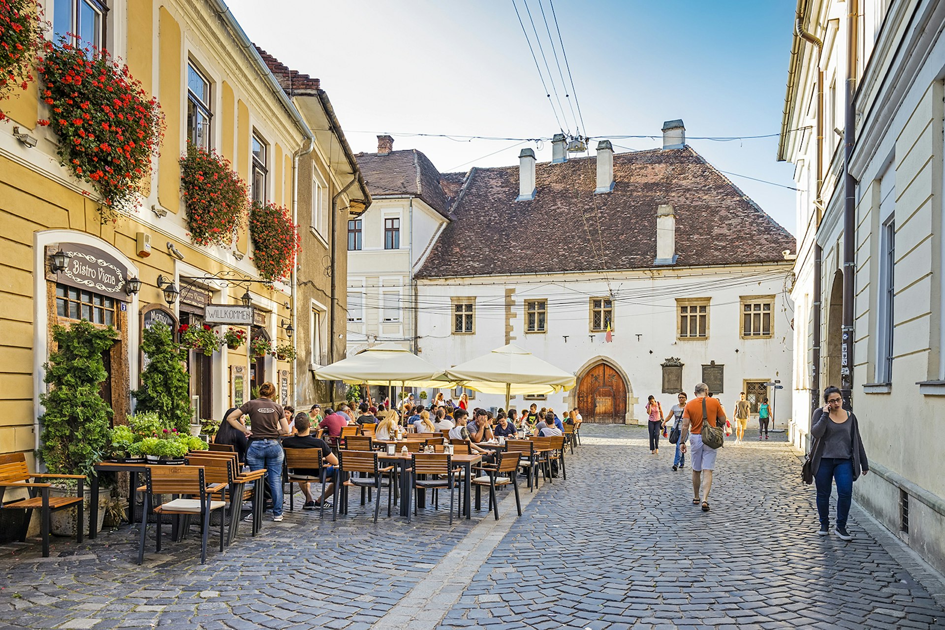 People sit on an outdoor patio in a cobblestoned plaza, surrounded by white and yellow historic buildings. The building on the left has large flower boxes in the windows. Cluj-Napoca, Romania.