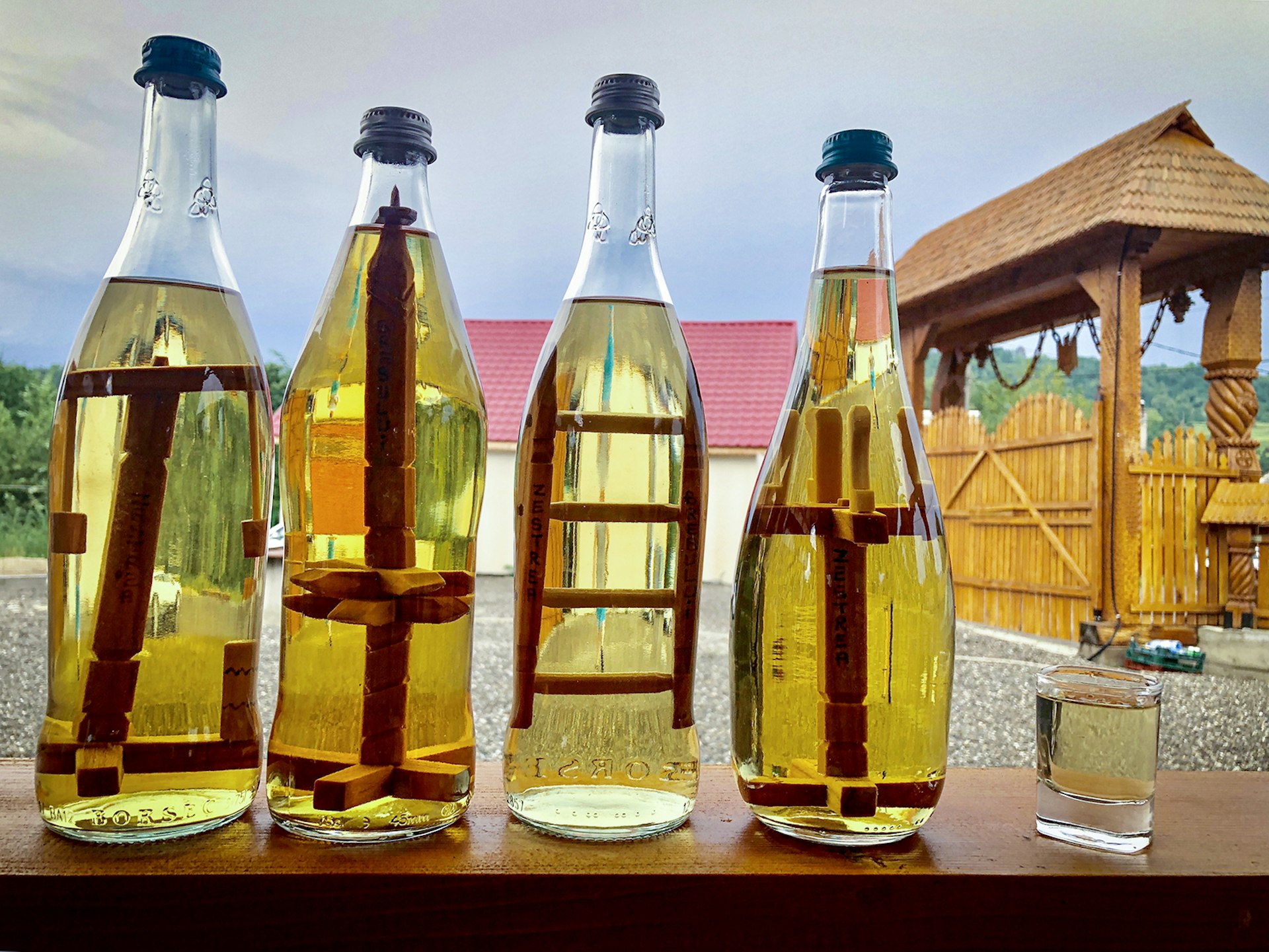 Three bottles of yellow liquor sit on a table next to a tasting glass with a barn in the background. Transylvania, Romania.