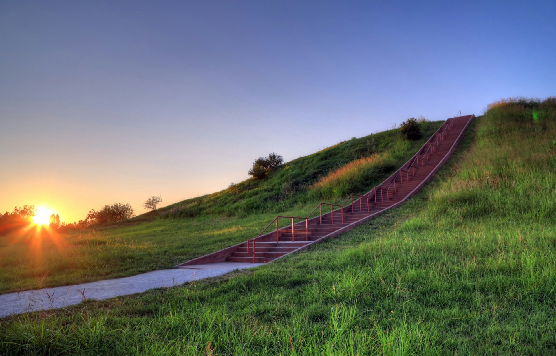 The sun sets behind a grassy mound with a staircase going up it. 