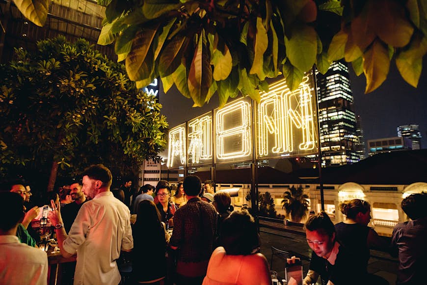 A crowd of patrons drinking and chatting at Loof bar below a neon bar sign, with glimpses of Singapore skyscrapers behind
