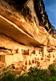 A stormy sky swirls above a cliff face with adobe buildings nestled inside.