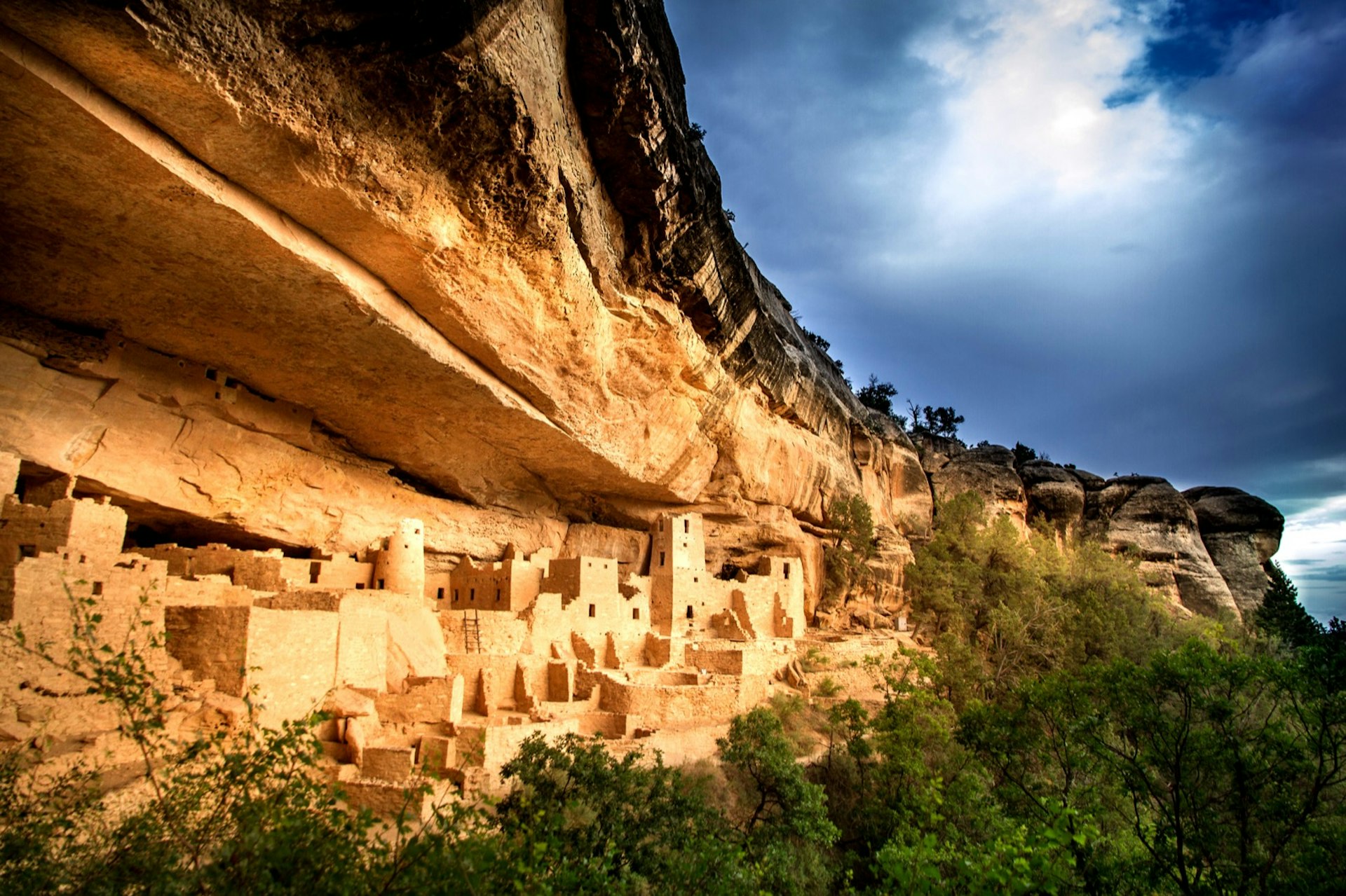 A stormy sky swirls above a cliff face with adobe buildings nestled inside. 