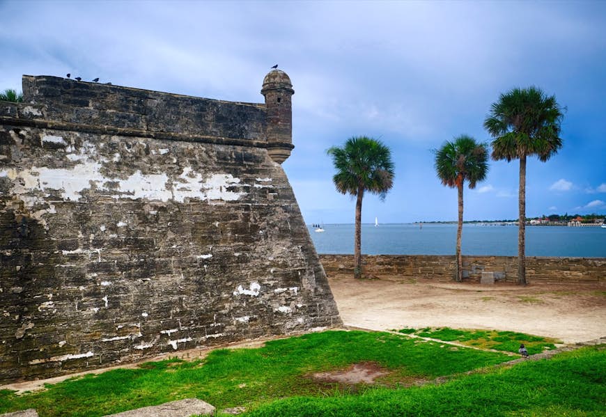 A stone fort looks out on the ocean as palm trees sway in the adjacent plaza