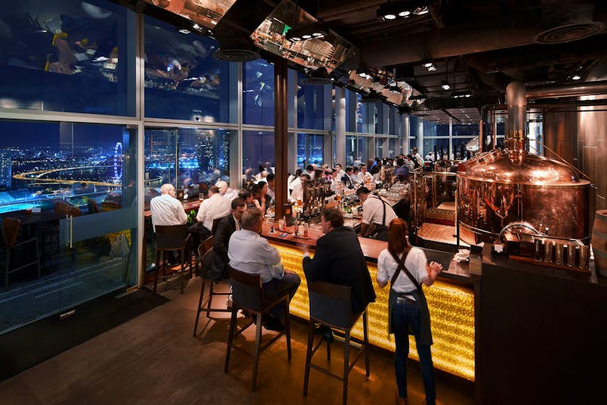 Patrons sit around a long bar next to large windows overlooking the skyline at night; a large brew-house kettle stands behind the bar © Level 33