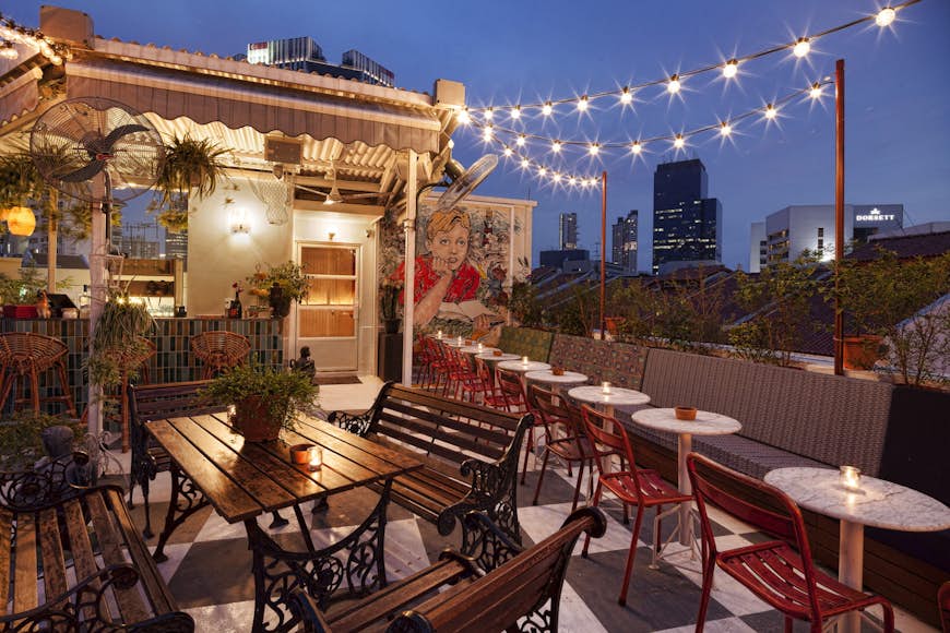 A small rooftop space with strings of lights above tables and chairs, some buildings of the skyline in the background © Potato Head Singapore 