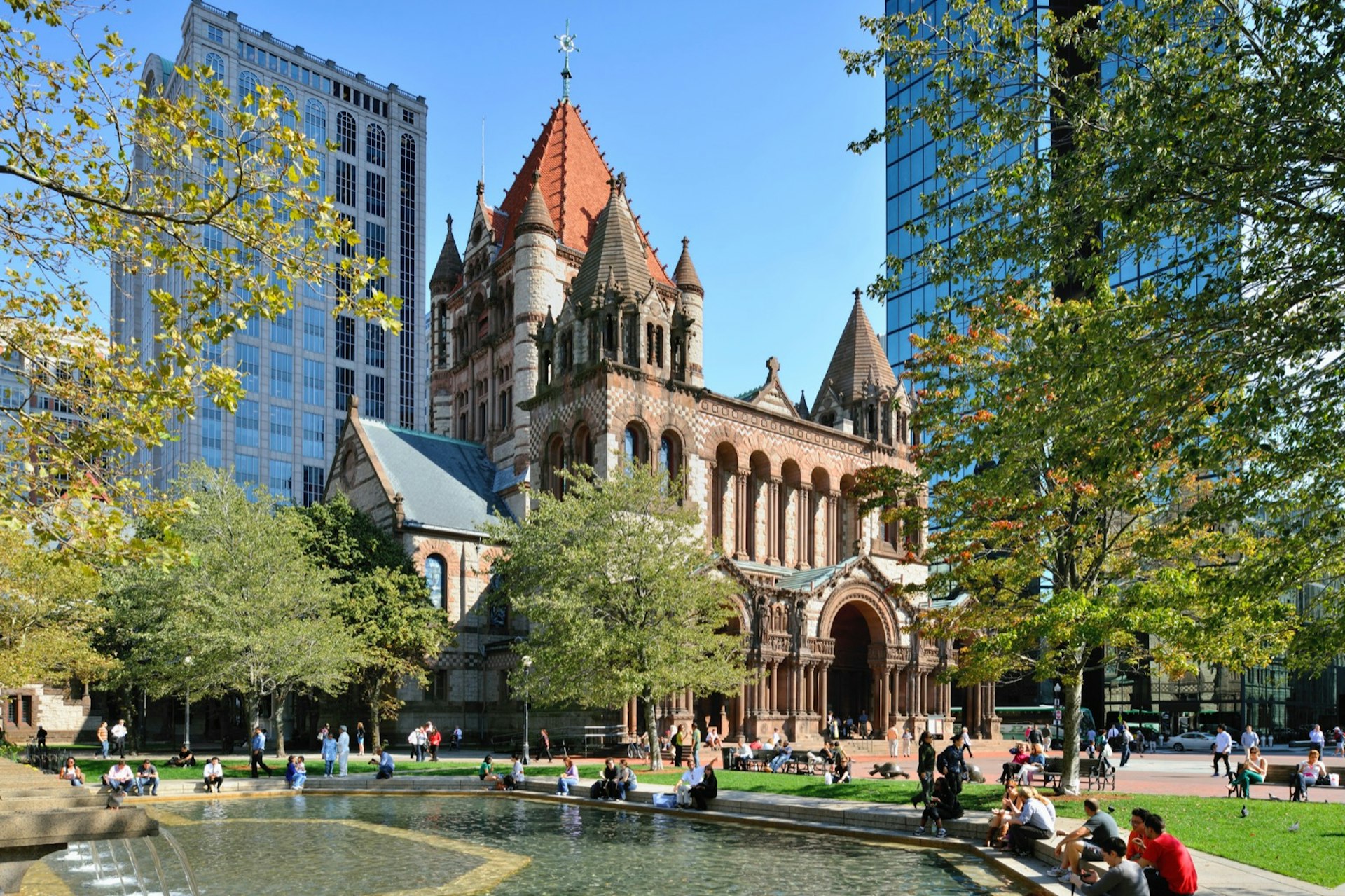 A group of people sit around a fountain and in front of Trinity Church on a sunny day in Boston
