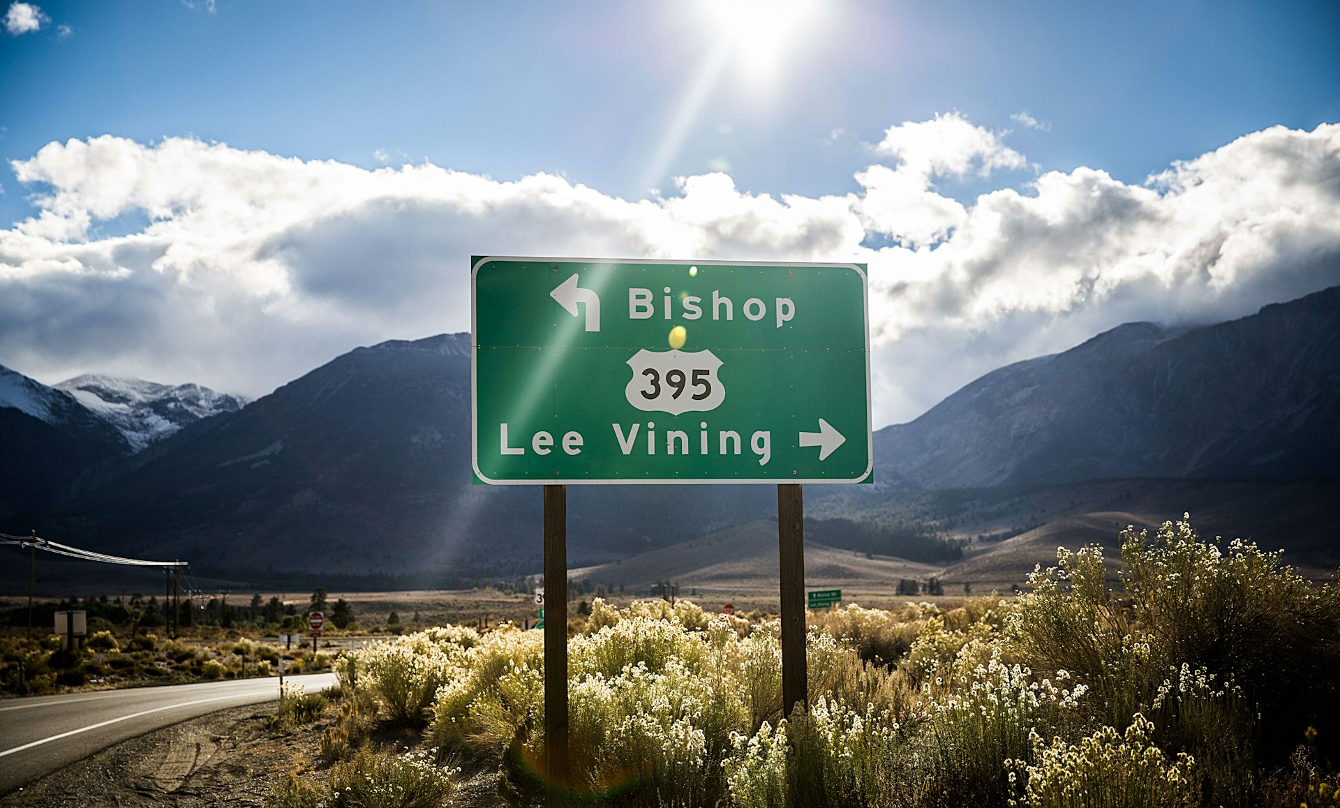 The sun shines behind a green road sign designating Hwy 395 in California and pointing to Bishop to the left and Lee Vining to the right.