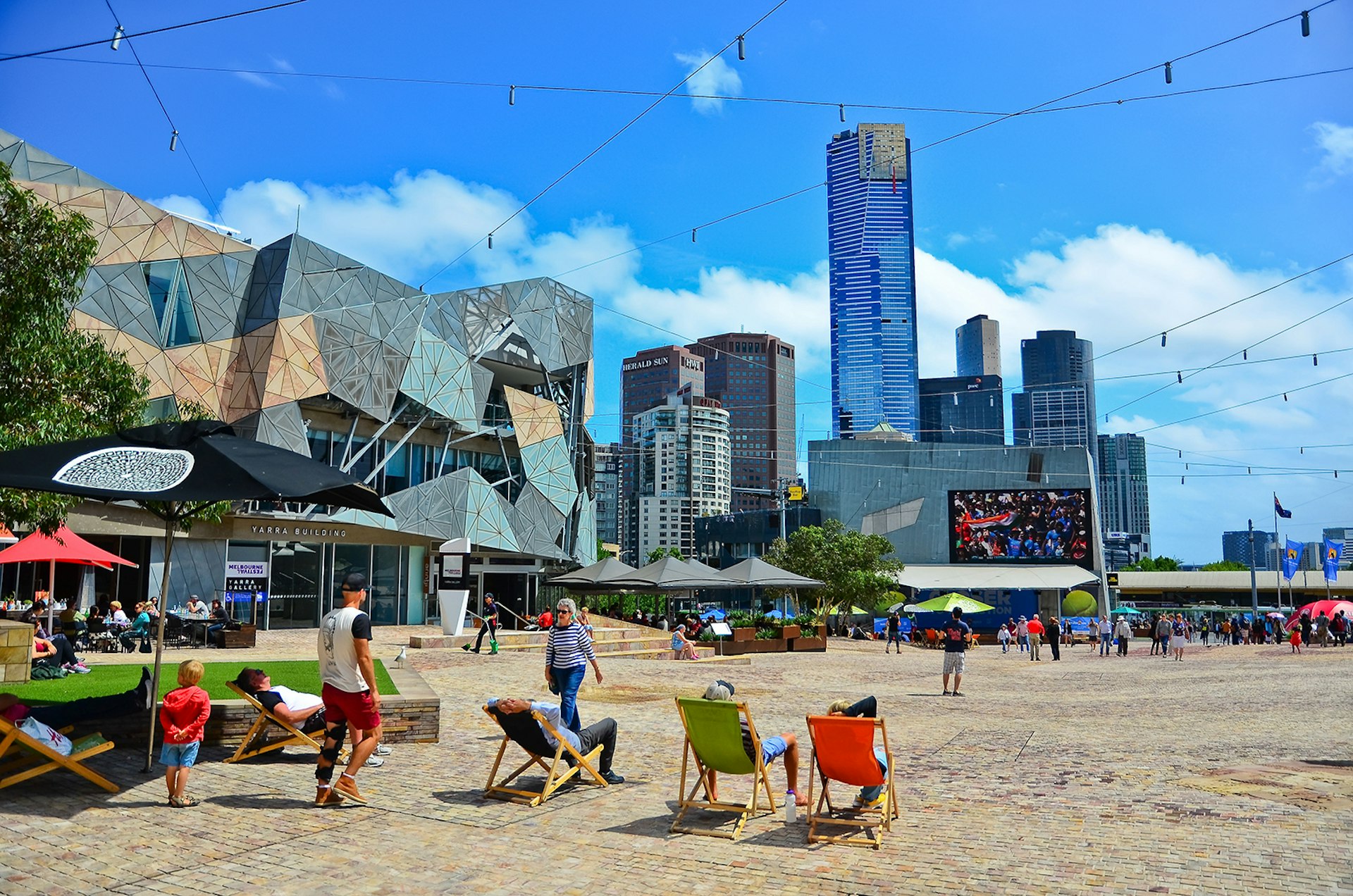 Tourists sit in multicolor sun chairs in Federation Square. A modern architecture building is visible as well as a skyline of business towers. Melbourne, Australia.
