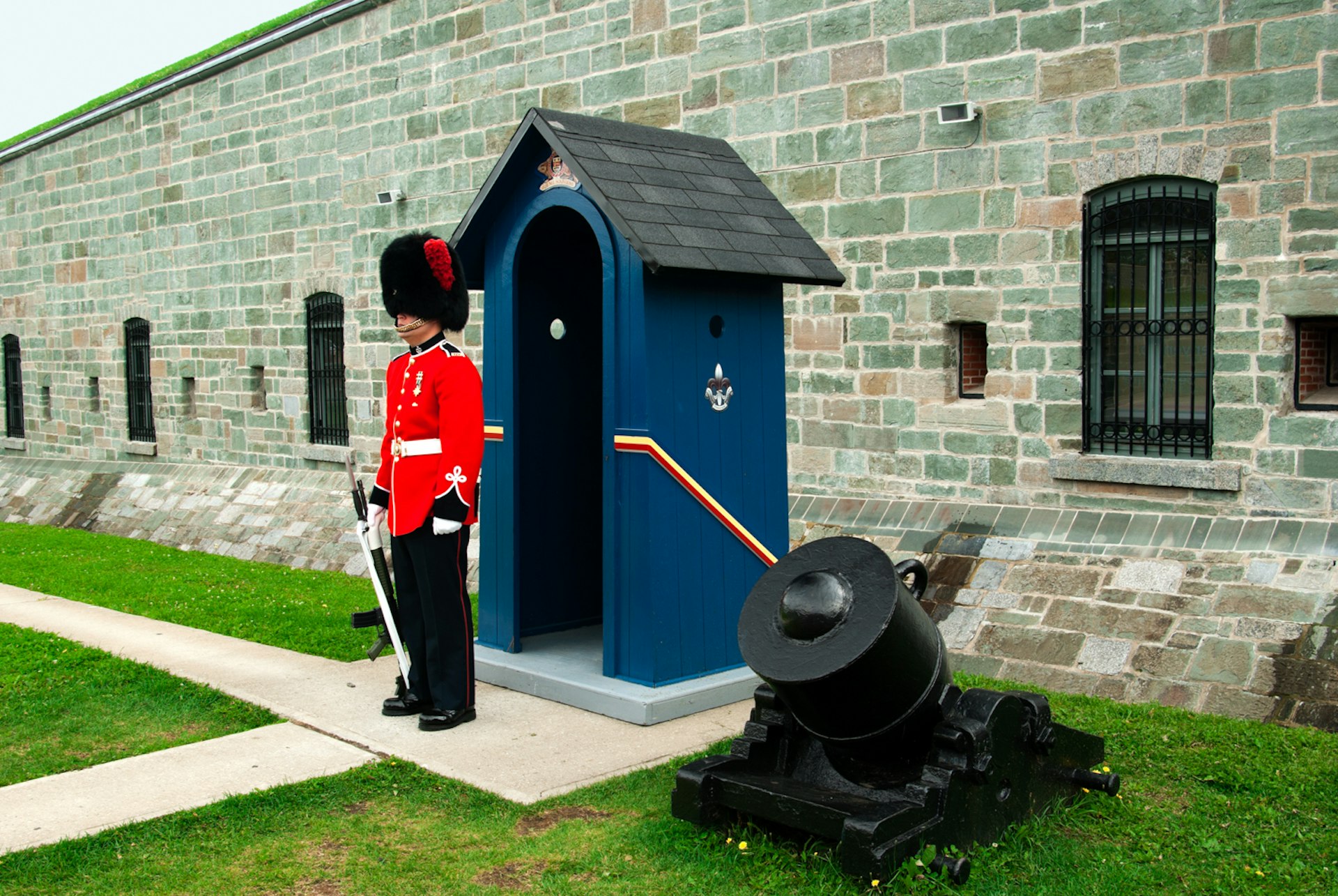 A guard with a sword in a red uniform and beaver hat stands watch at the Citadelle outside a blue shed