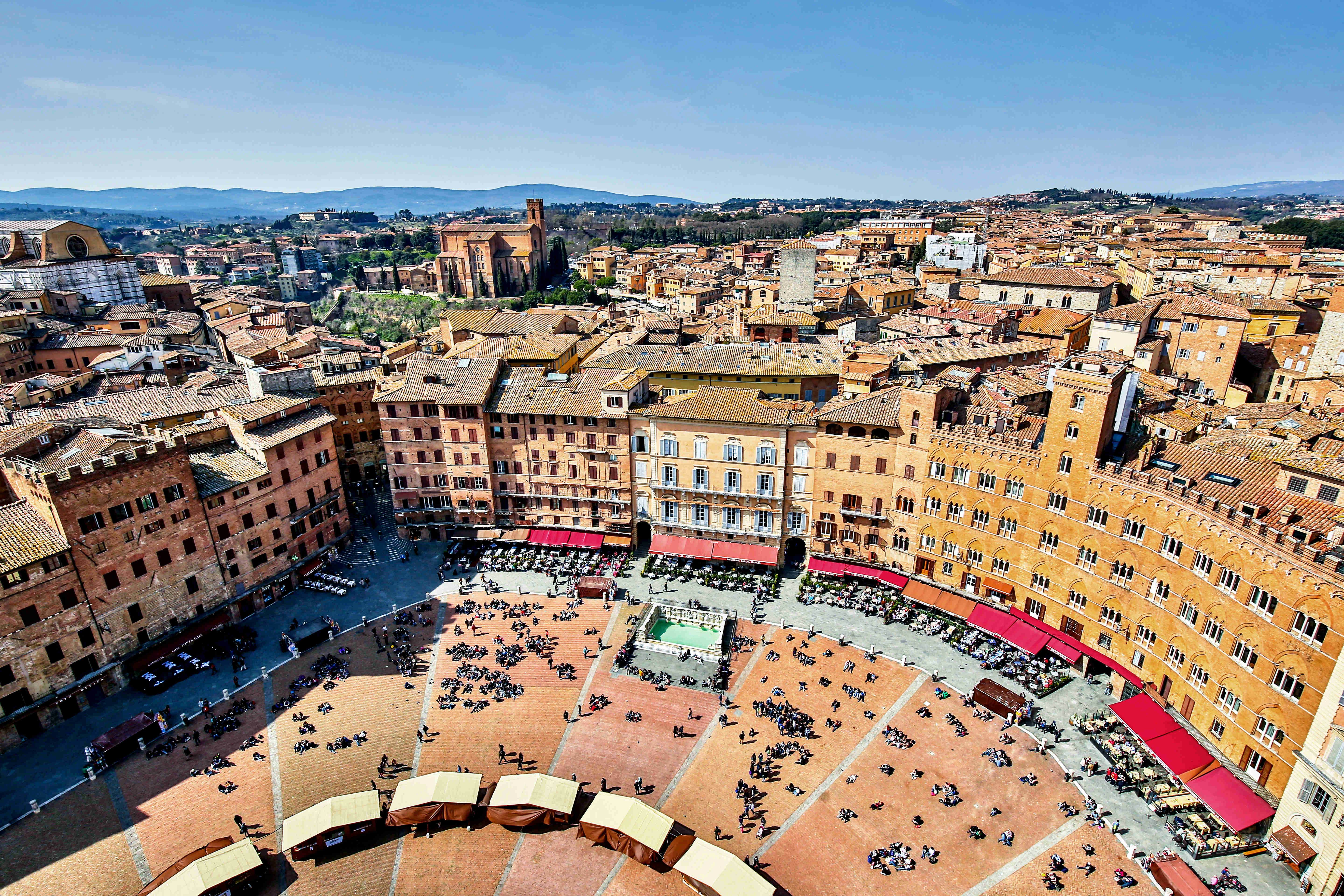 A high overhead photo shows people milling about a red-and-orange hued piazza surrounded by tall stone buildings.