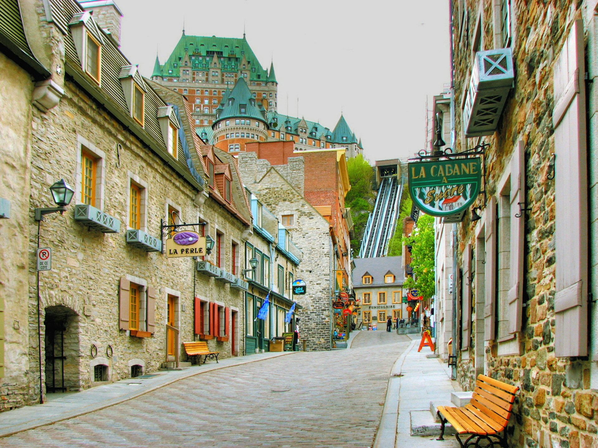 Québec City’s cobblestone streets with stone buildings either side that have been turned into various shops and cafes. Château Frontenac, a large red brick building with a copper roof and turrets, is visible in the background. 