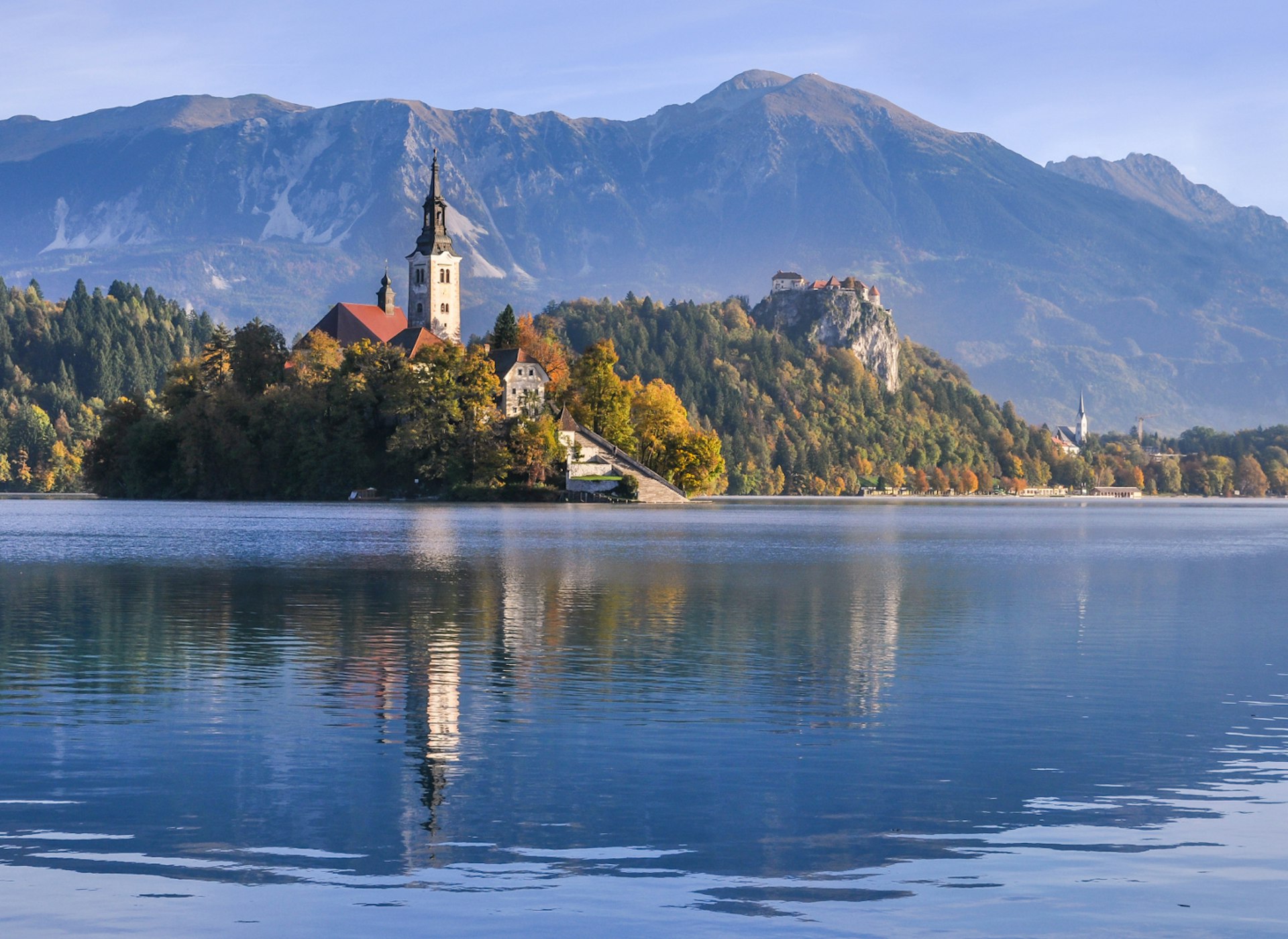 A church on an island in the middle of a lake. A castle sits on a rocky outcrop behind the island, and there are mountains in the distance.