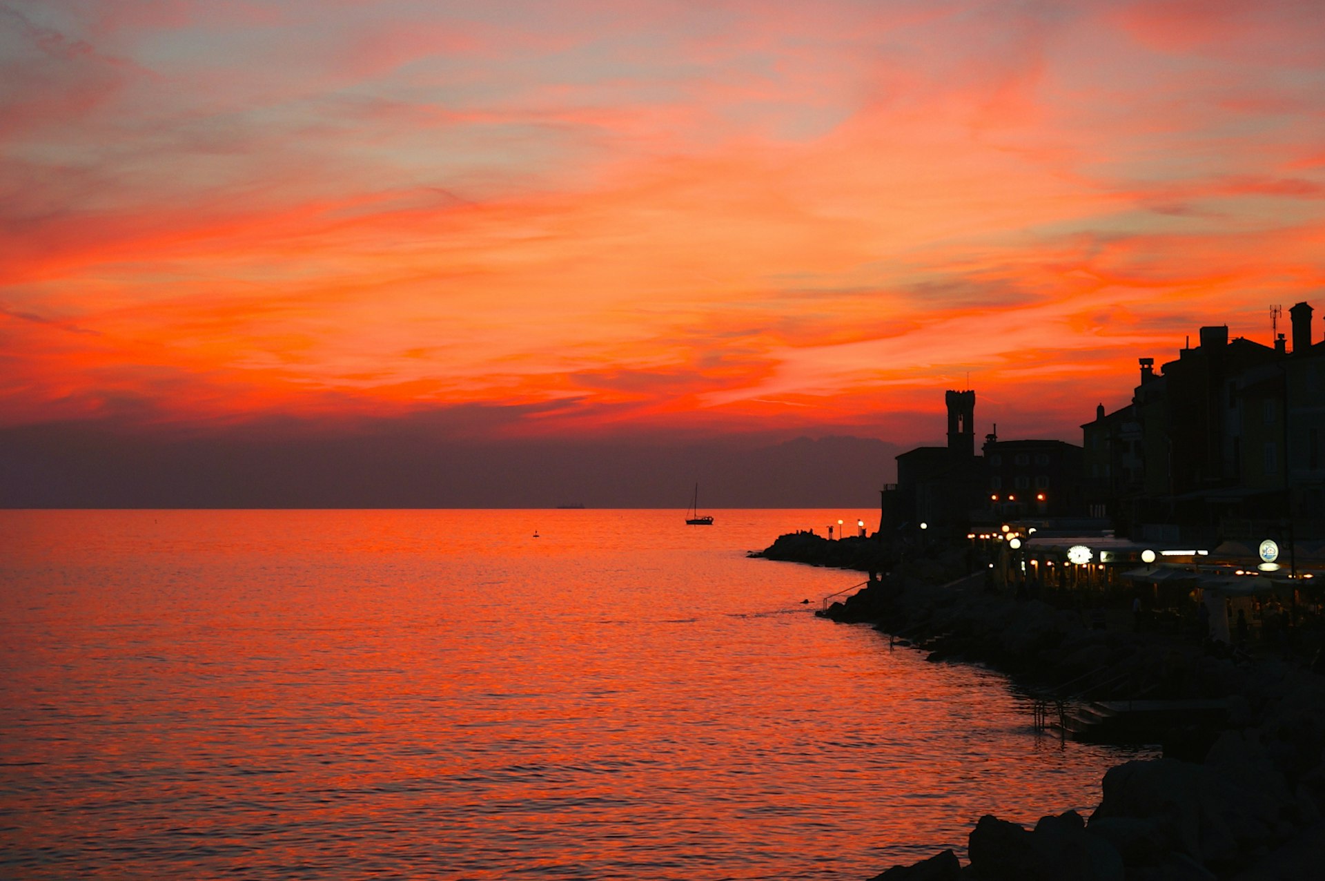 An orange and purple sky is reflected in the calm sea. Buildings lining the shore and a boat in the sea appear as silhouettes