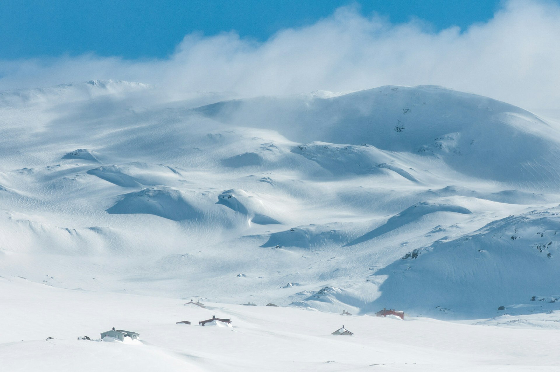 Snow drifts off the top of a range of snow-covered mountains. At the base are small lodgings nestled in the snow