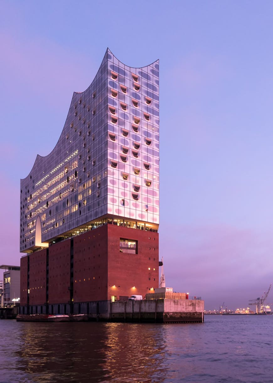 The Elbphilharmonie, viewed from the river, at dusk. A pink ligh is reflecting on the building's mirrored surface 
