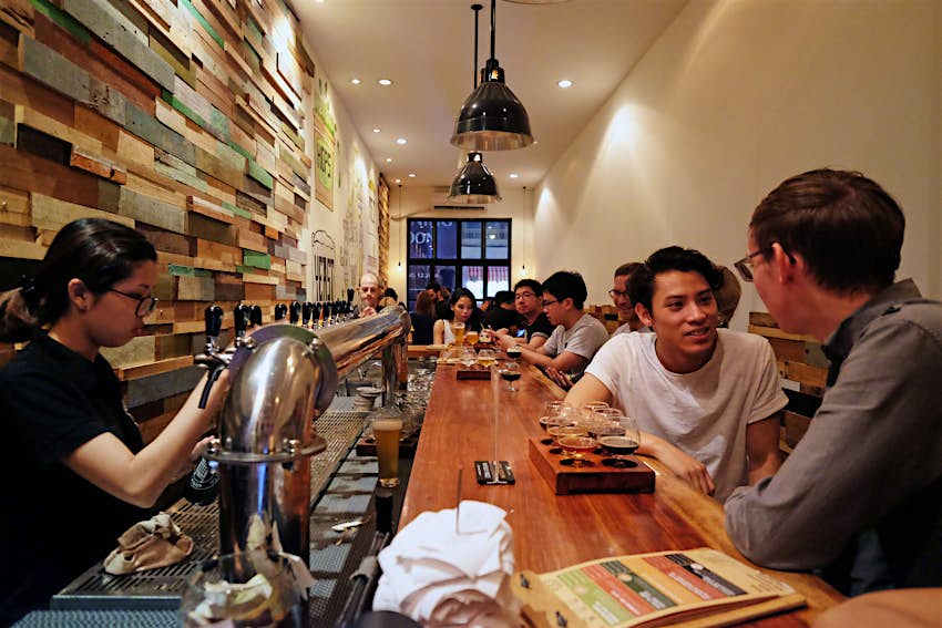 Patrons sit and drink at a long narrow bar while a server pours a beer at Pasteur Street Brewing Company