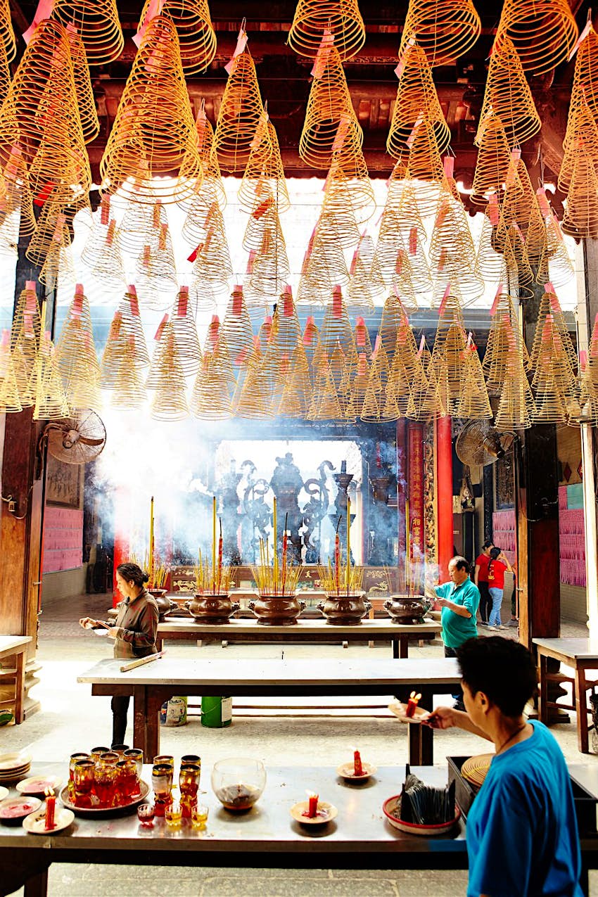 Features - Candles and incense in Thien Hau Temple. Large cone-shaped coils of incense hang from the ceiling of Thien Hau Pagoda, incense sticks give off smoke, while a visitor lights a candle below