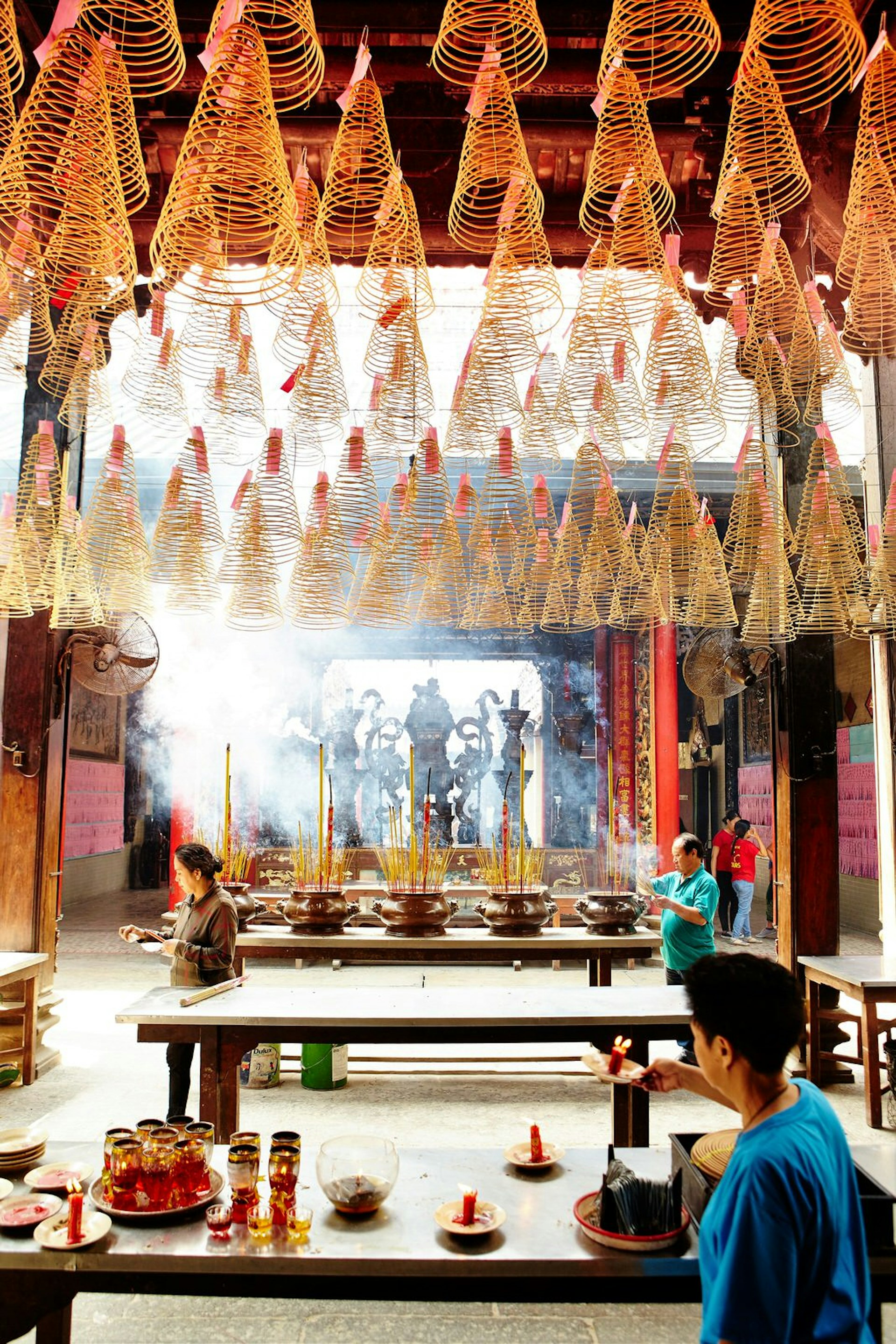 Large cone-shaped coils of incense hang from the ceiling of Thien Hau Pagoda, incense sticks give off smoke, while a visitor lights a candle below