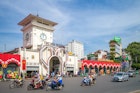 The entrance to the market is a white facade with a clock tower that stands tall against a blue sky. Several motorcycles speed by in the road in front of the market building