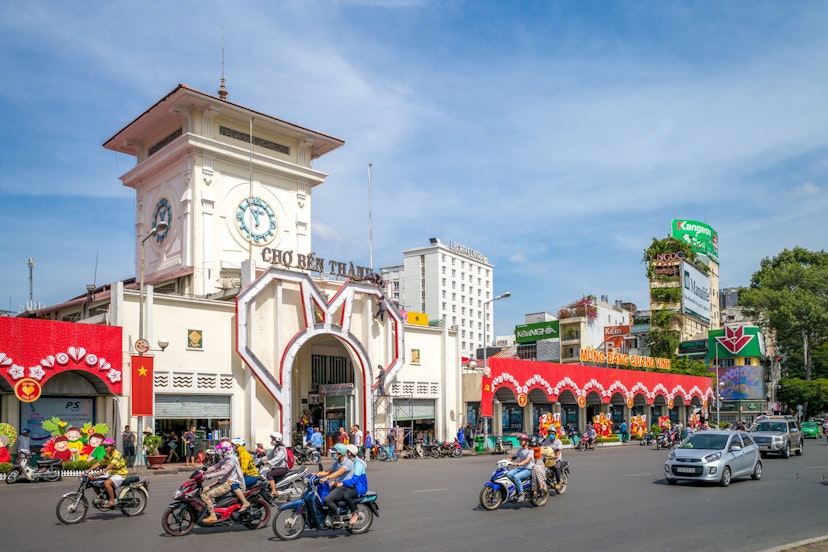 The entrance to the market is a white facade with a clock tower that stands tall against a blue sky. Several motorcycles speed by in the road in front of the market building