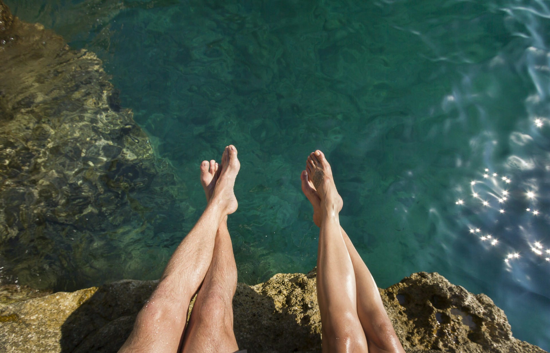 A view from above of two people's legs over a rock face with the sea below