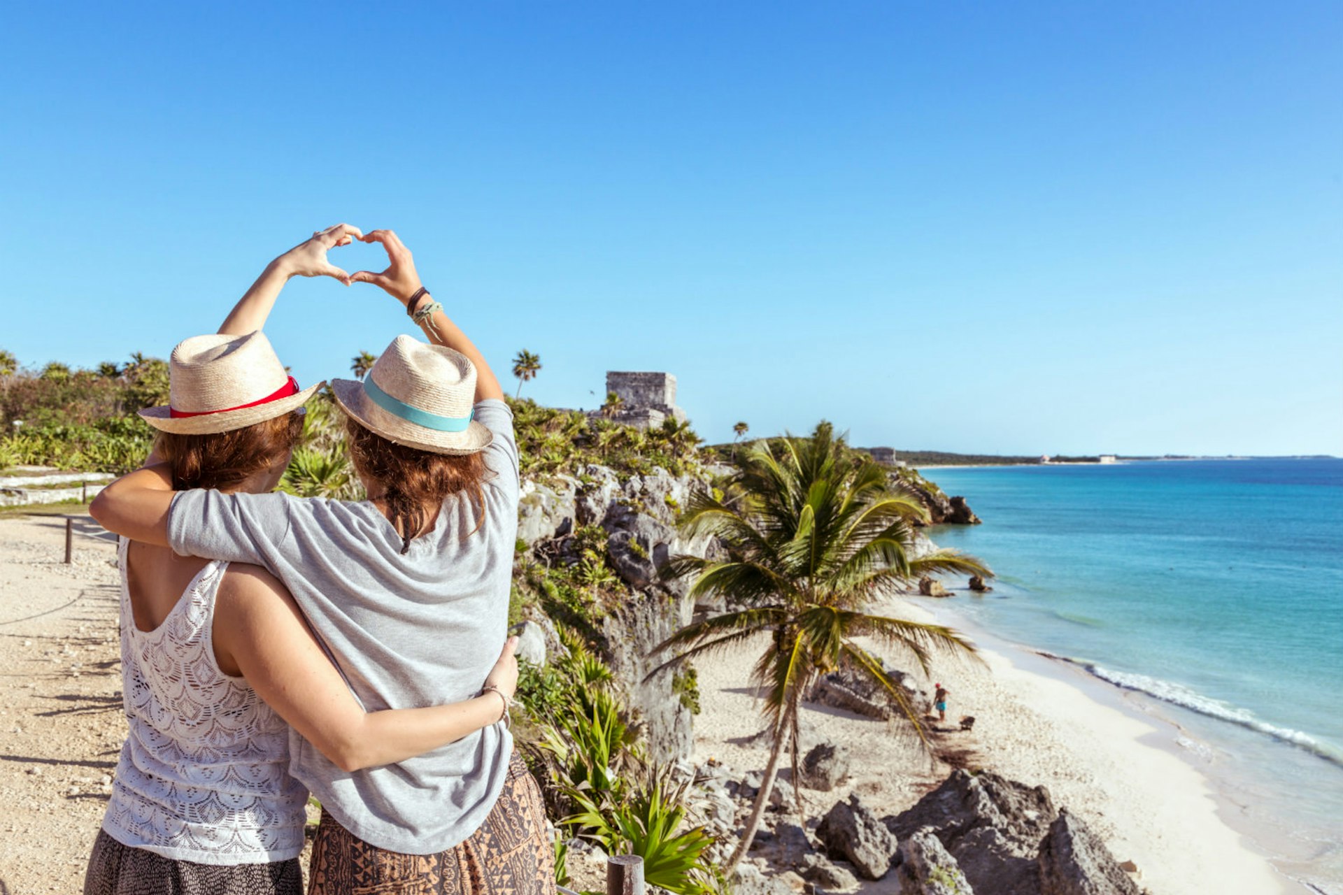 An embracing female couple form a love heart with their hands in front of a tropical beach