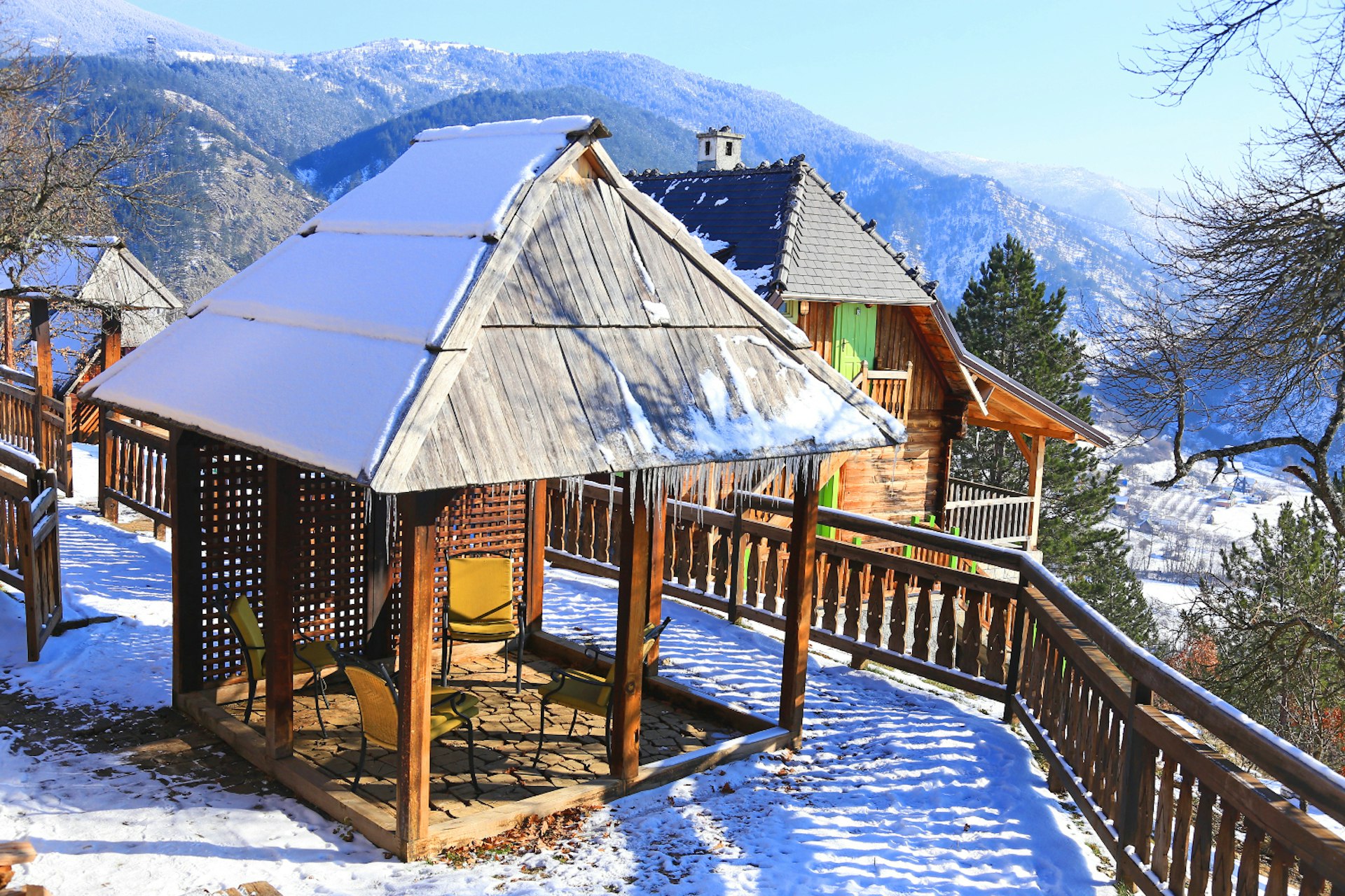 An open-sided wooden hut sits on a snowy hillside. A larger wooden chalet is in the background