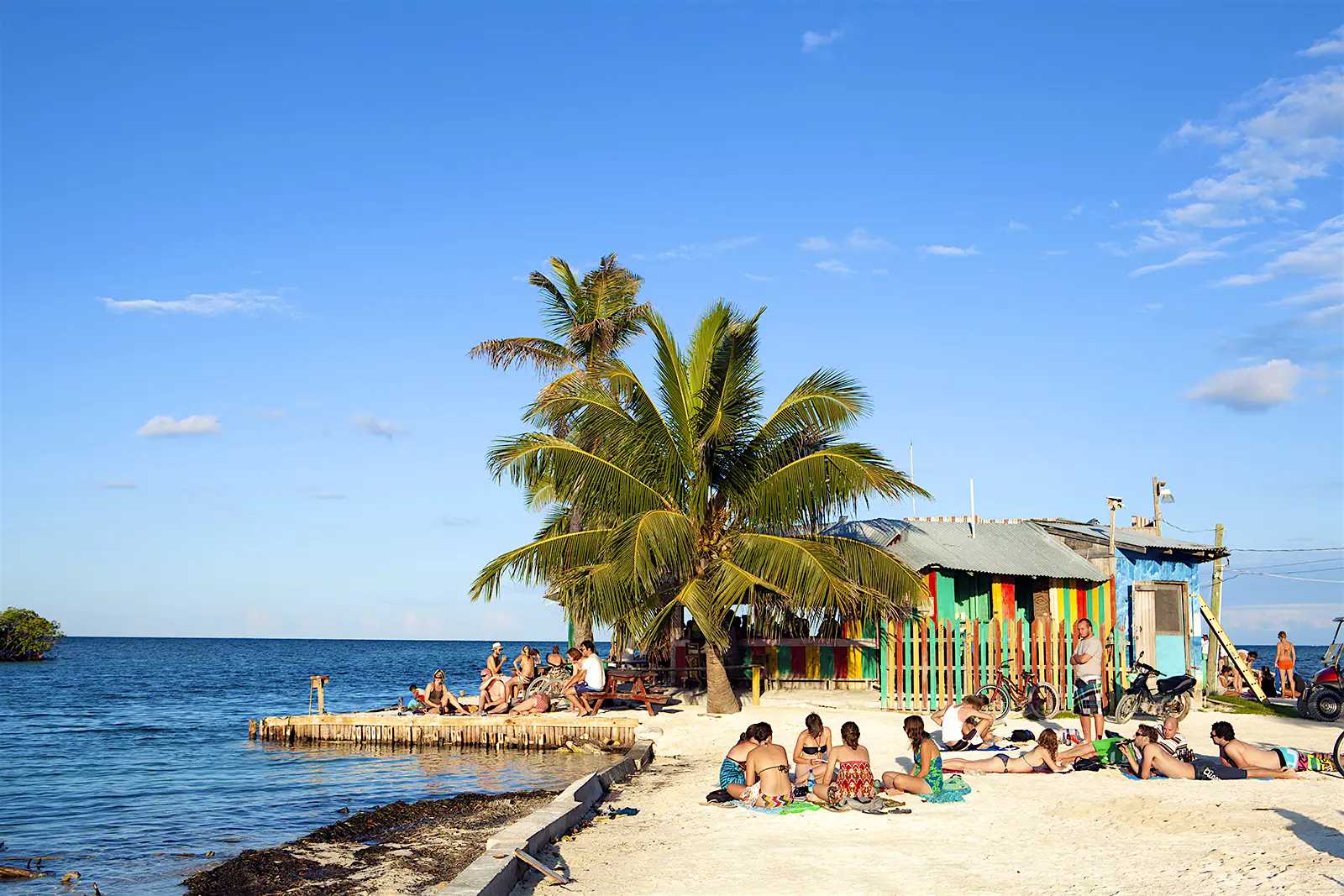 Features - Central America, Belize, Belize district, Caye Caulker, tourists sit on the tiny beach under a coconut palm outside the Lazy Lizard bar at the split between the two halves of the island People sit on the small beach outside the Lazy Lizard bar © Alex Robinson / Getty Images