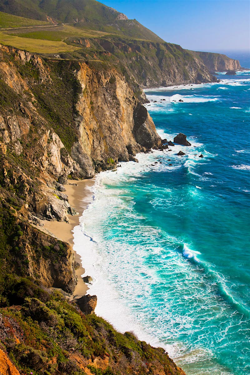 Features - shutterstock_134985383-c6f061ff7bc0 Big Sur's coastline. Cliffs with shrubbery and exposed rock face plunge down to meet a rough, blue sea. A road winds along the top of the cliff.