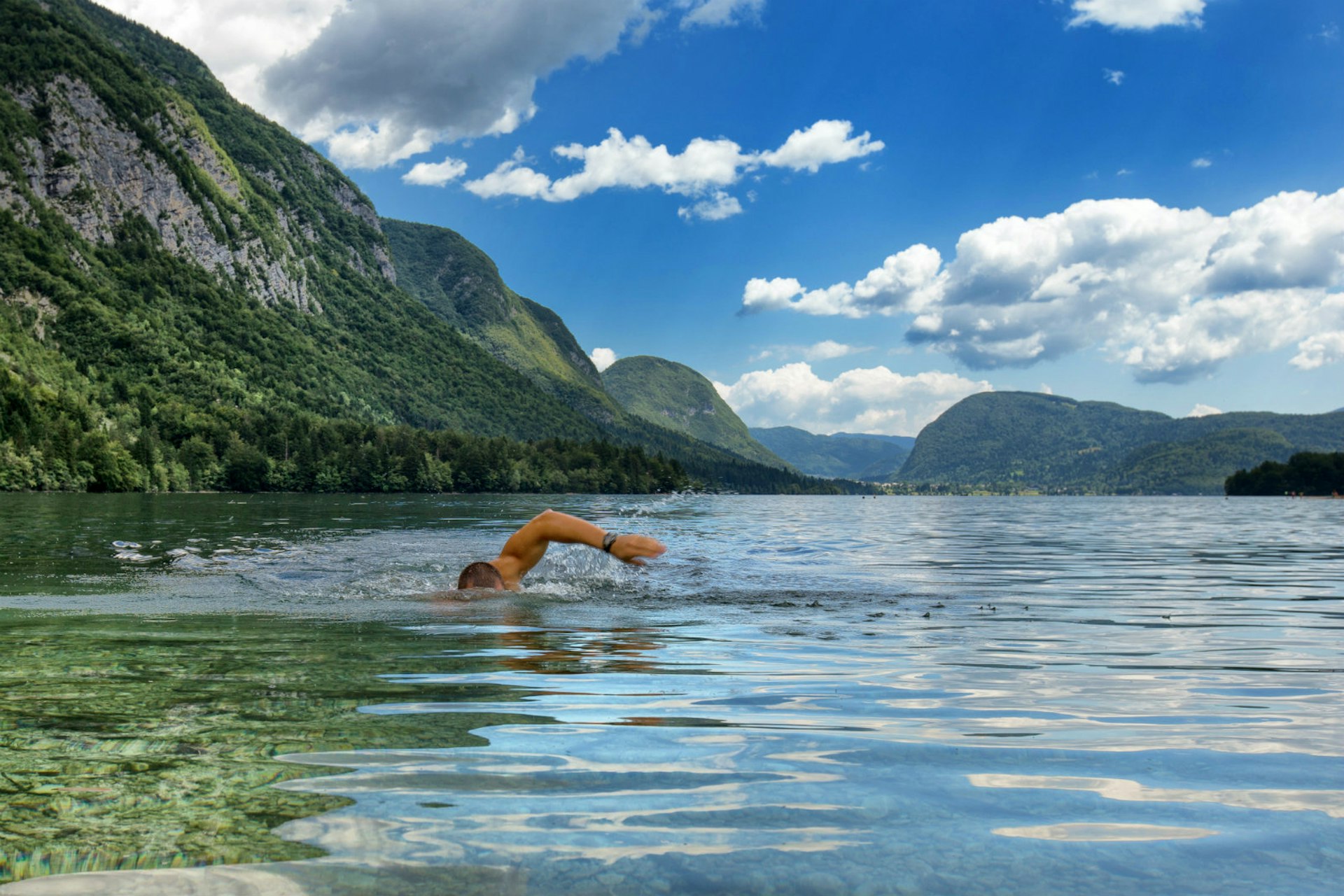A man swims through the waters of Lake Bohinj, with mountains in the background and a cloudy blue sky overhead