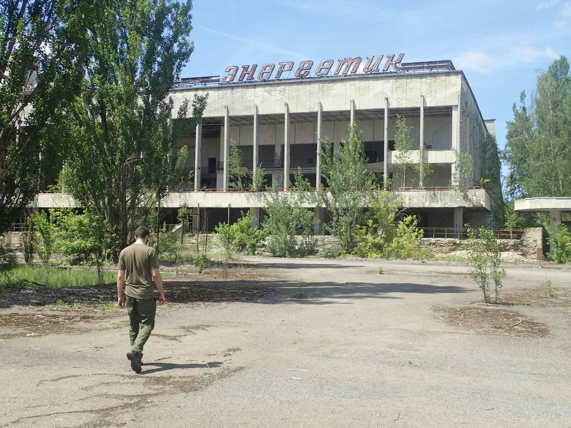 A guide walks across tarmac towards the derelict Palace of Culture in Pripyat; tress are growing in front of and within the pillared concrete structure, which has a large sign in Ukrainian script on its roof. 