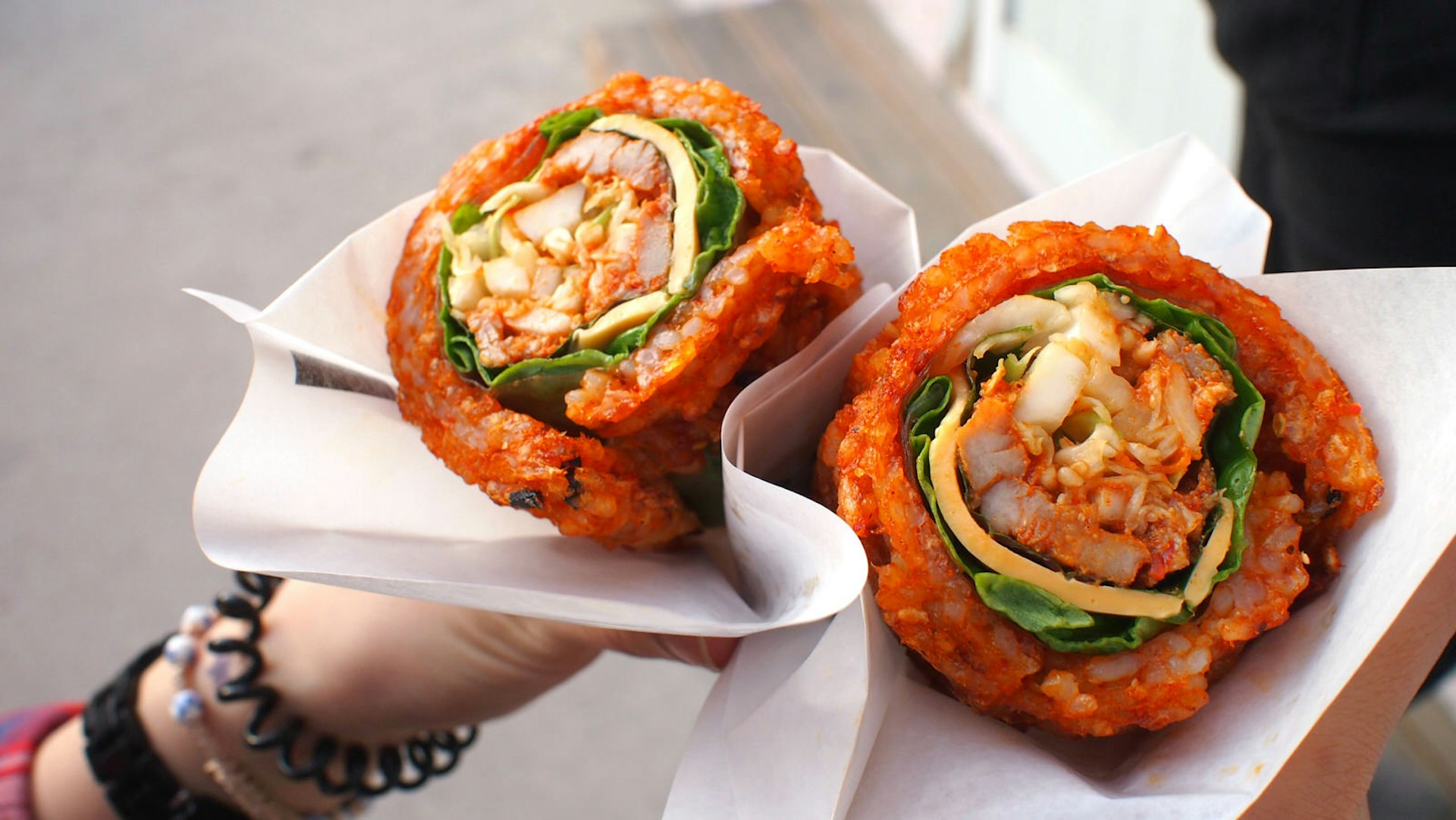 Bibimbap waffle is one of famous street food in Jeonju Hanok village, South Korea. It is composed of spicy rice rolled around a filling - pieces of meat, vegetables, cheese, and rice paper. A woman wearing a bracelet is holding it in her hands.