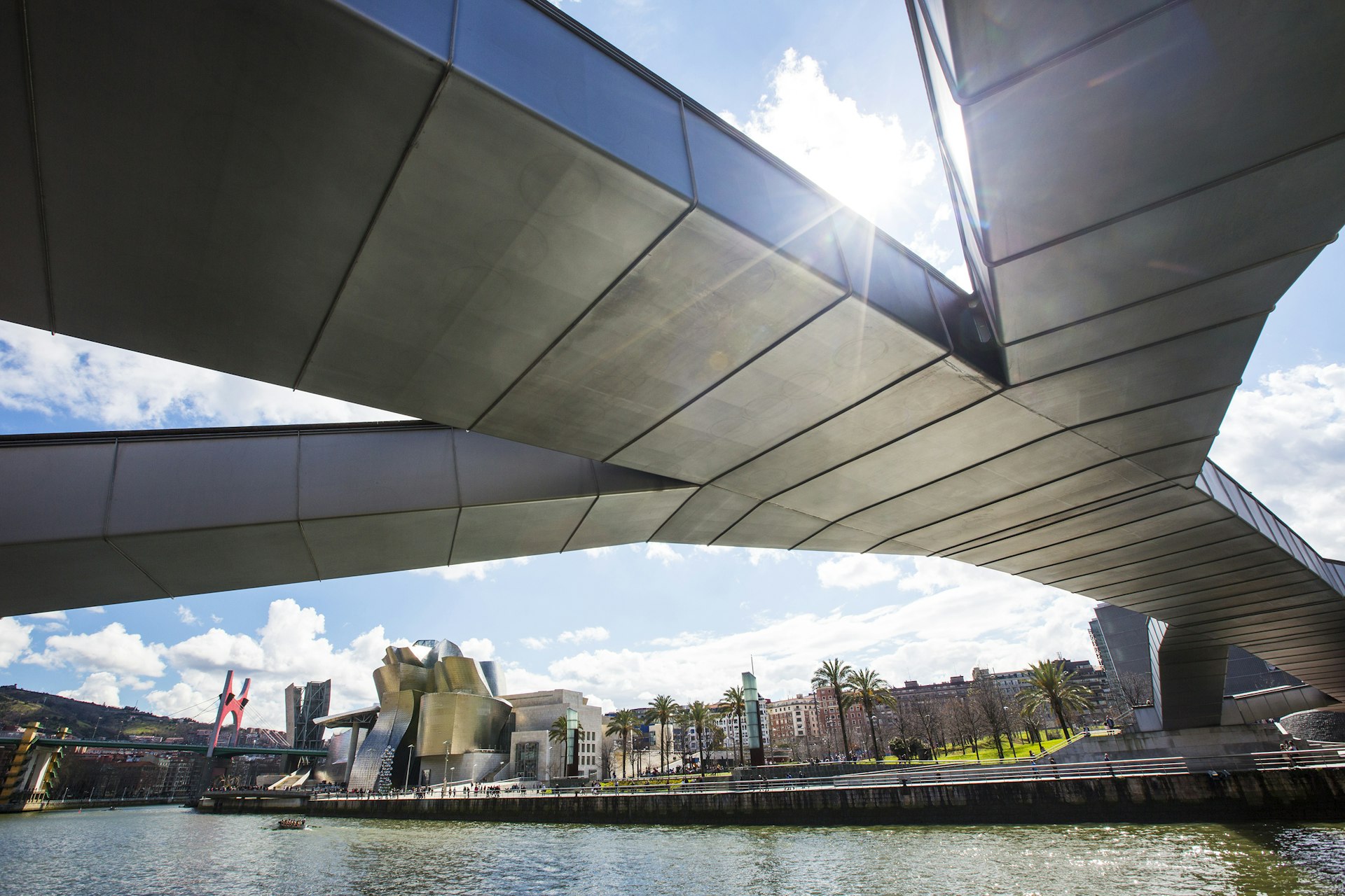 An image of a silver footbridge from below with a view of the gleaming Guggenheim on the banks of Rio Nervión in the background; visit Basque country