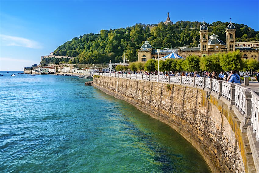 Features - La Concha bay in San Sebastian A group of people walk across a bridge in San Sebastián. There's a towering structure and large trees in the background; visit Basque country