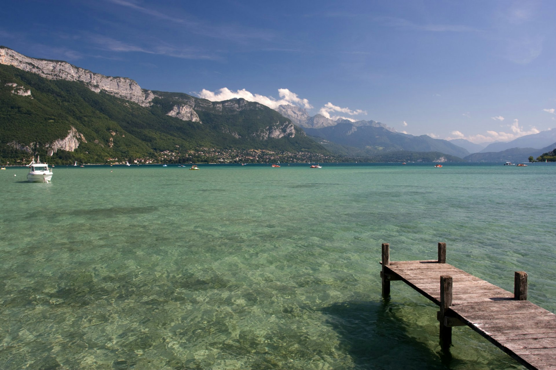 A large, still lake, with a settlement across the water at the foot of a mountain. Several boats are on the lake, and a wooden pier leads out into the water