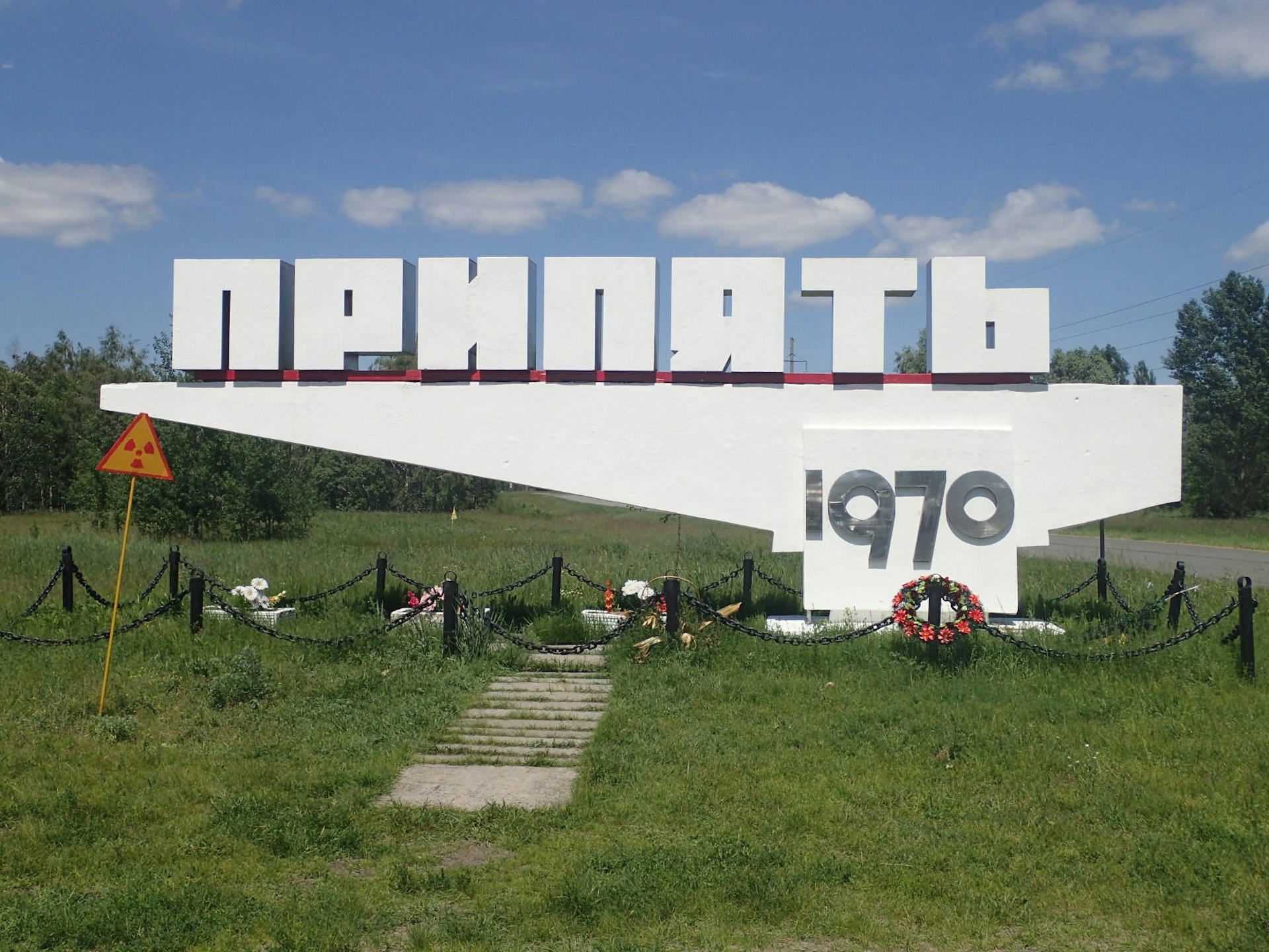 A concrete sign in Ukrainian script that translates as "Pripyat 1970", when the city was founded; wreaths and flowers are arrayed beneath it and a radiation warning sign stands next to it. 