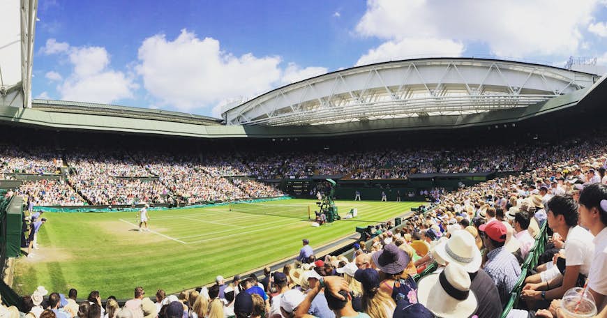 Sun pours down into Centre Court with the open roof exposing the players and spectators to a blue sky; shot from the corner courtside, the view look across the court, with Novak Djokovic serving to Kei Nishikori on the opposite side