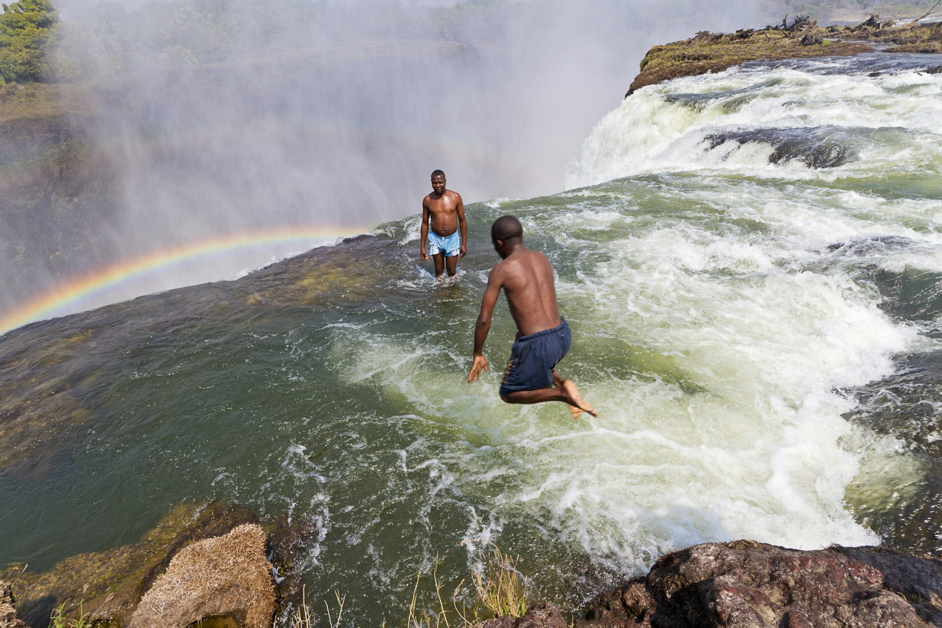 Man jumps into Devil's Pool at Victoria Falls. It looks like she will be swept over the waterfall but a thick lip of rock keeps people safe. Victoria Falls is nearly a mile wide and 360 feet deep and from the air, looks like the earth has been ripped in two. Zambia, Africa.