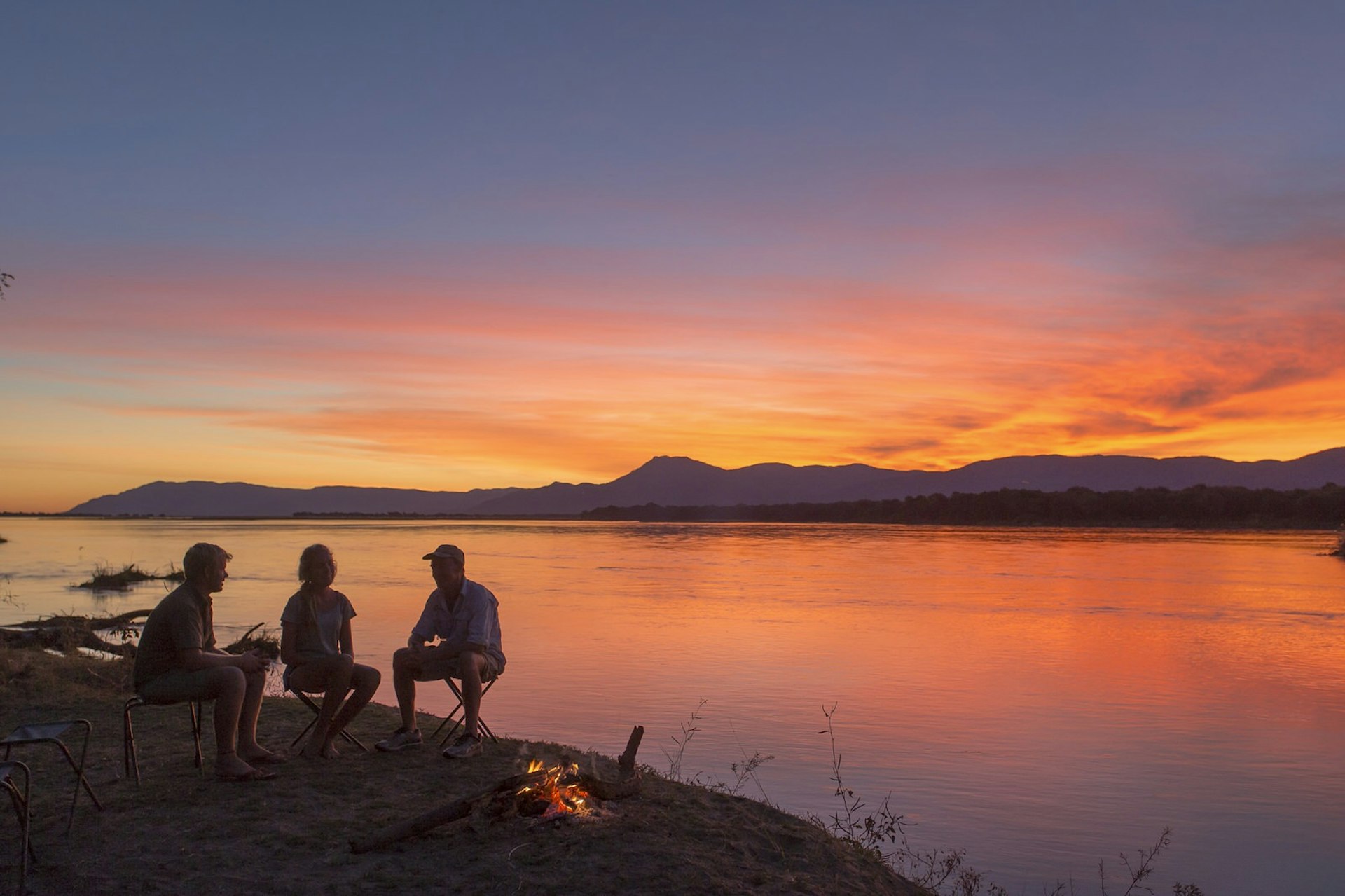 Three adults chat around a campfire on the banks of the Zambezi, Zambia © Philip Lee Harvey